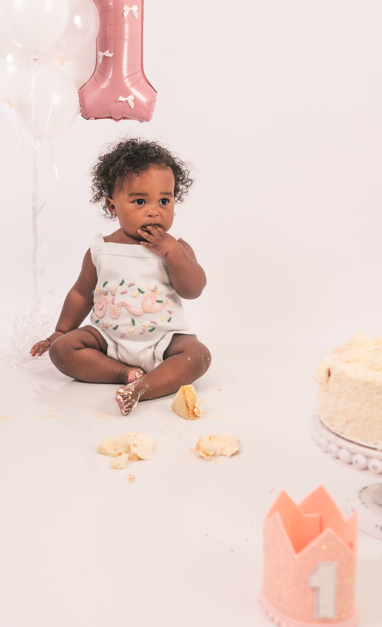 Baby in white floral romper sits on floor during first birthday photoshoot with pink decorations and cake.