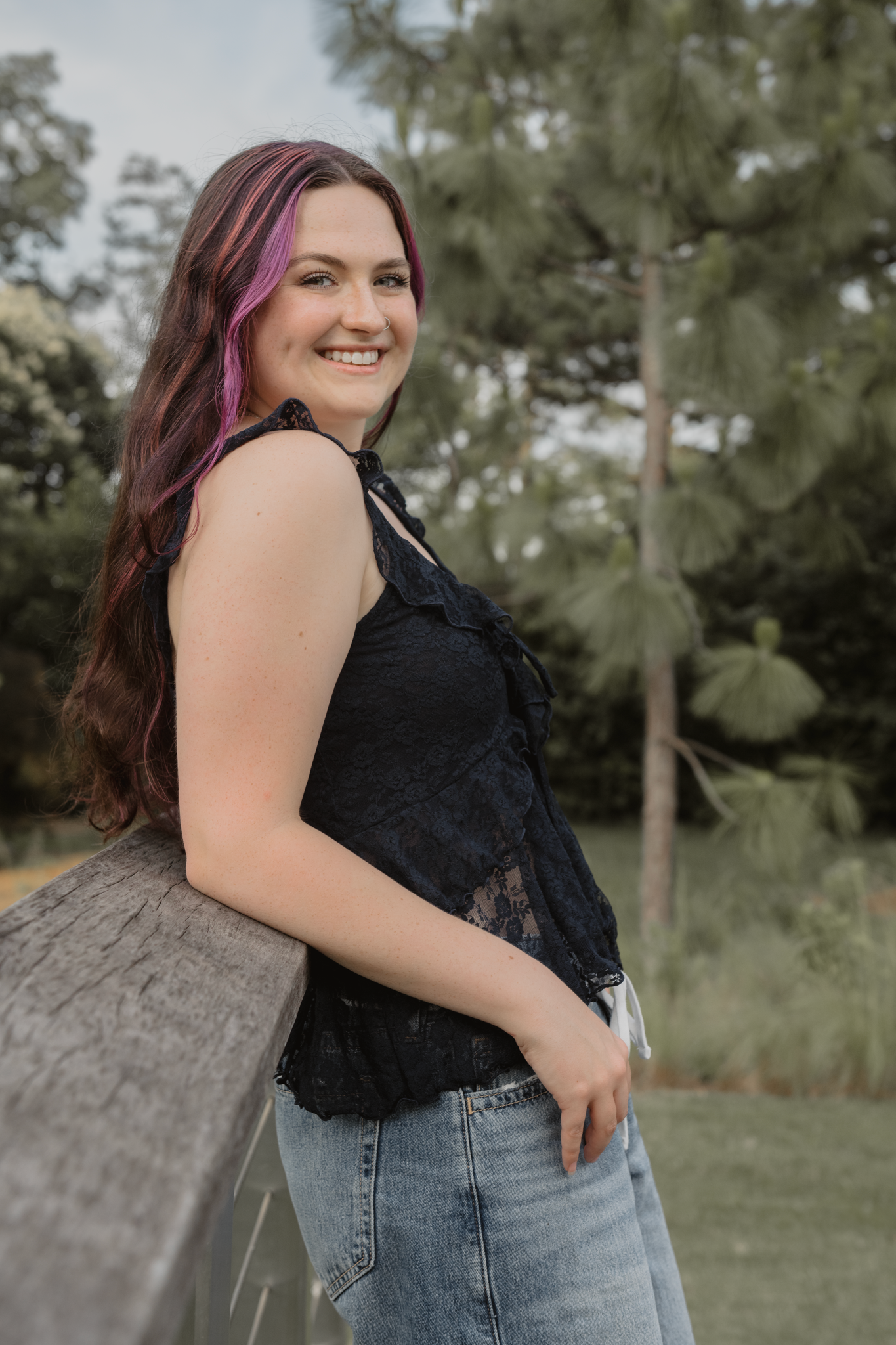 Haylee in a blue lace top and jeans poses casually against a wooden railing in an outdoor setting with trees during her senior session