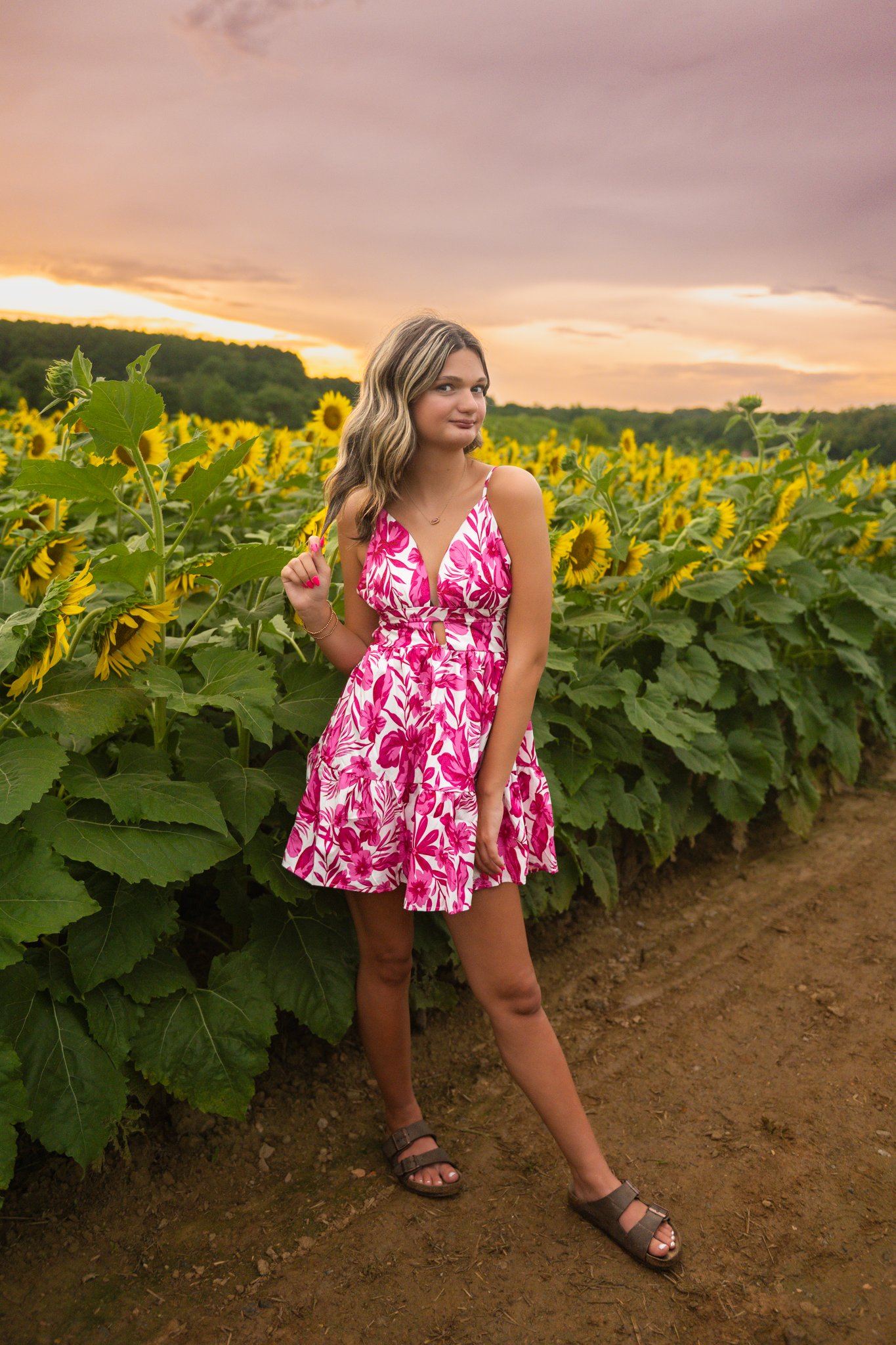 A person in a pink floral dress poses in front of blooming sunflower field during golden sunset hour.