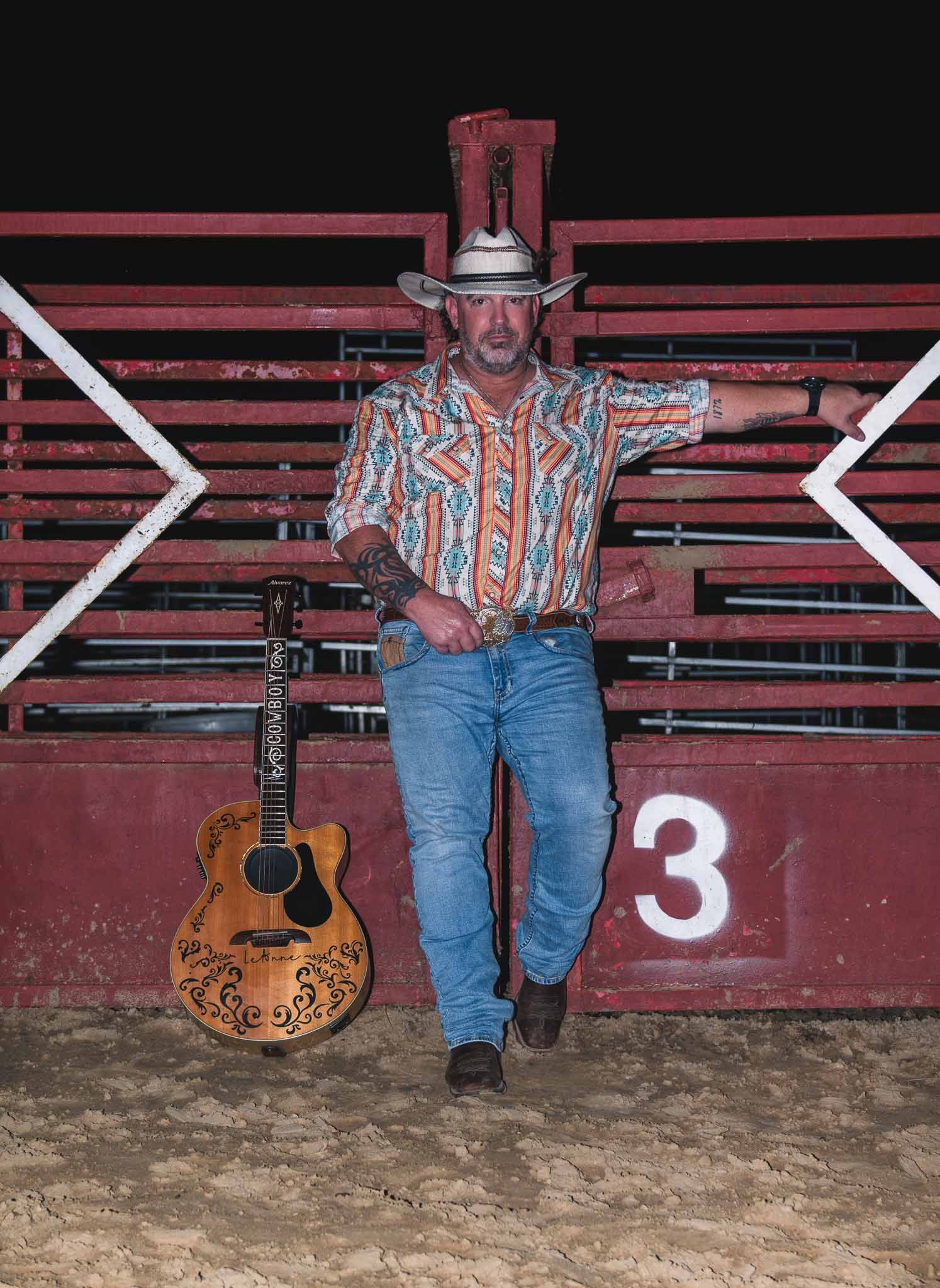 A country western performer in a striped shirt and cowboy hat poses against red bleachers with an acoustic guitar nearby.