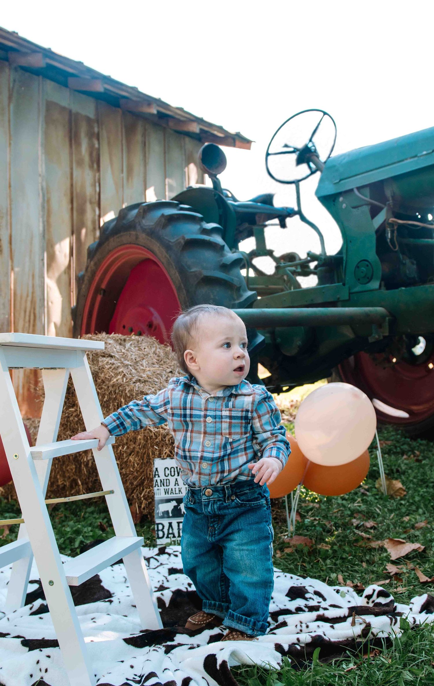 A toddler in plaid and denim stands by a ladder and hay bales with a vintage tractor in the background.