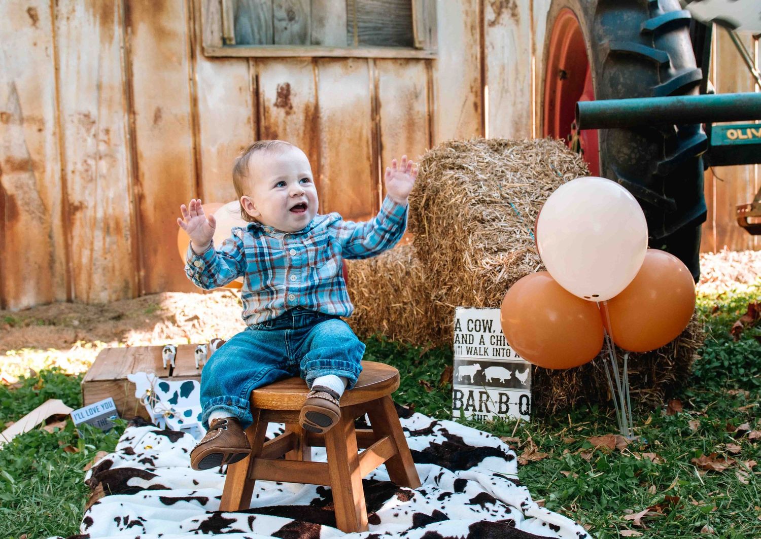 A small child sits on a wooden stool with balloons beside them at a rustic outdoor setting with a cow print blanket.