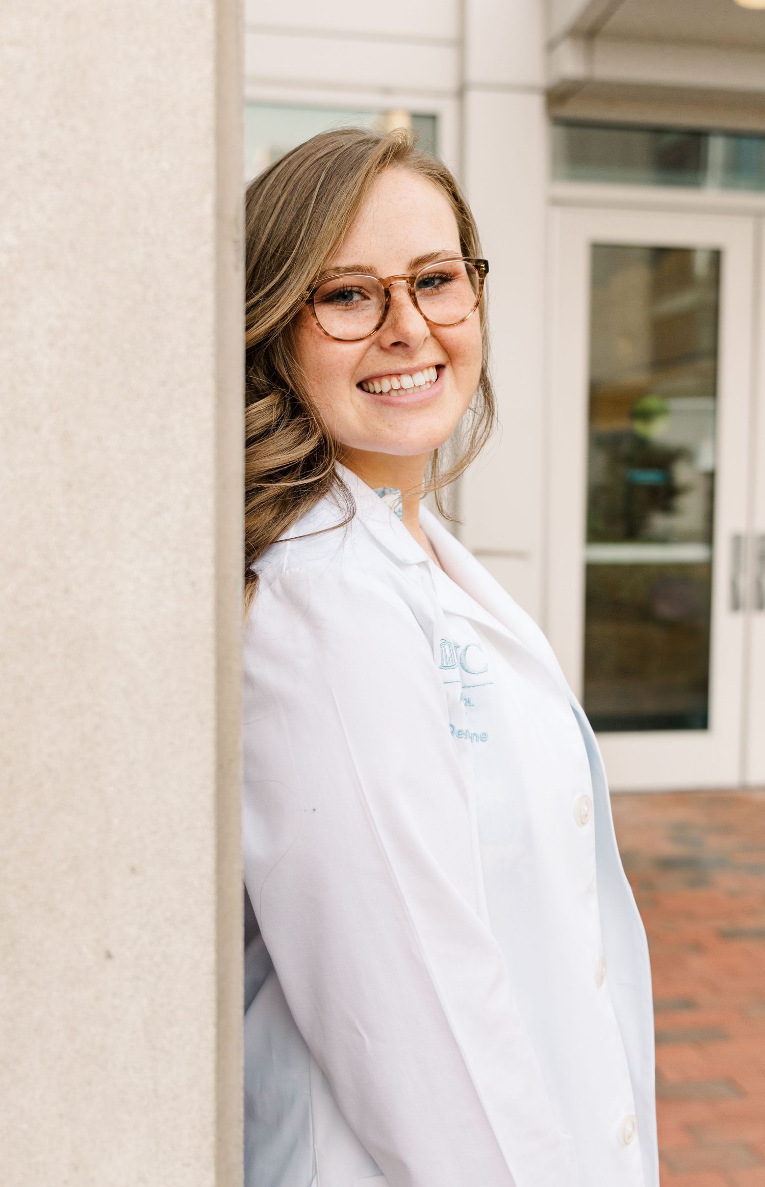 Healthcare professional in white coat and glasses smiles while leaning against a column outside a medical building.