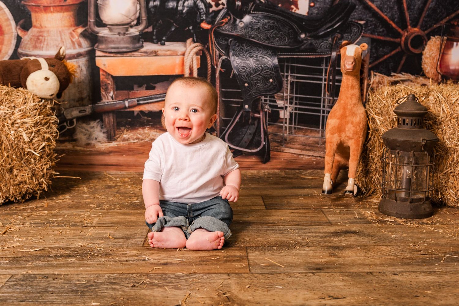 A baby sits on wooden floor surrounded by rustic farmhouse decor including hay bales and vintage items.