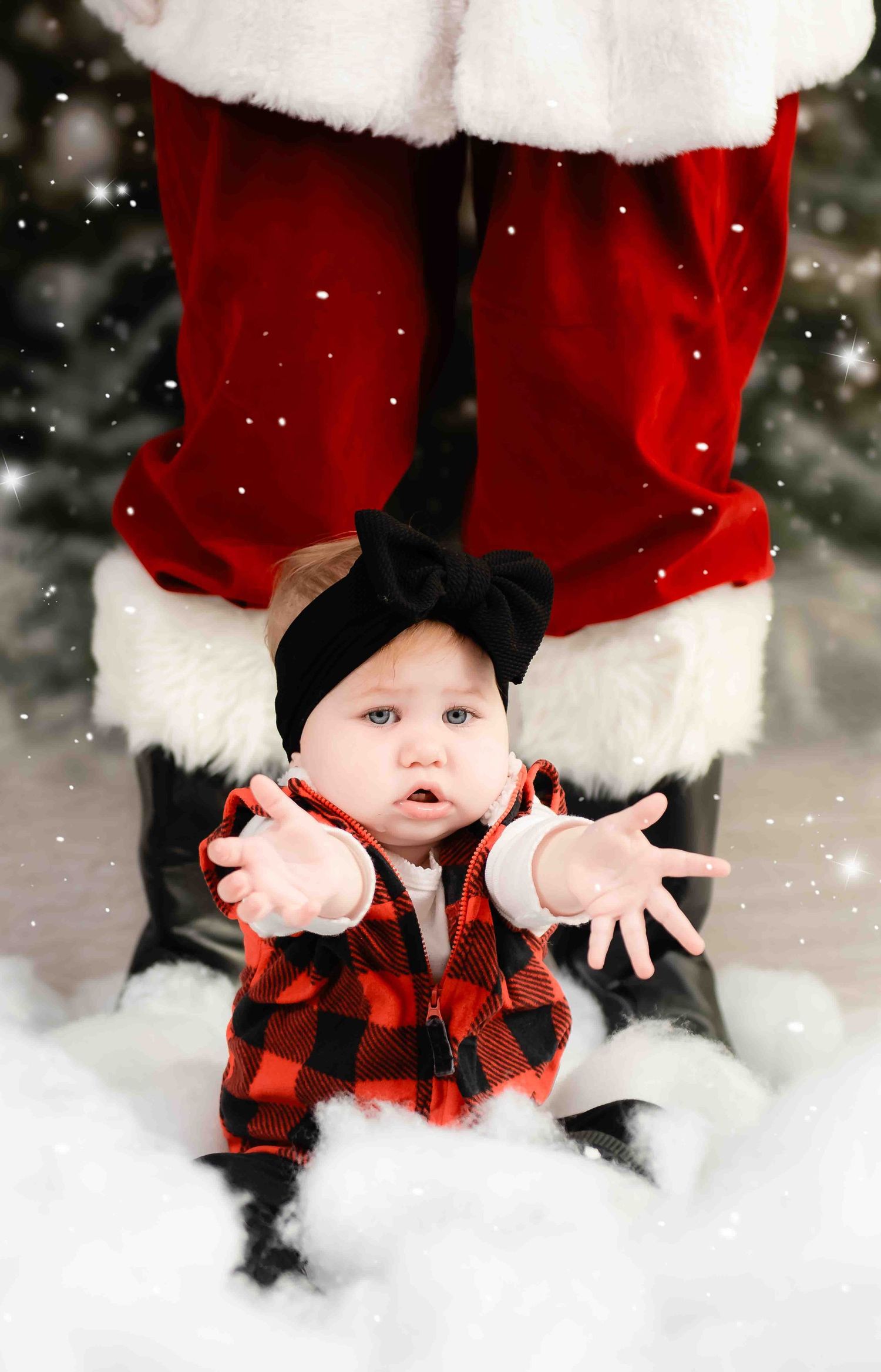 A baby wearing buffalo plaid and a black bow sitting in snow in front of Santa's red pants and white trim.
