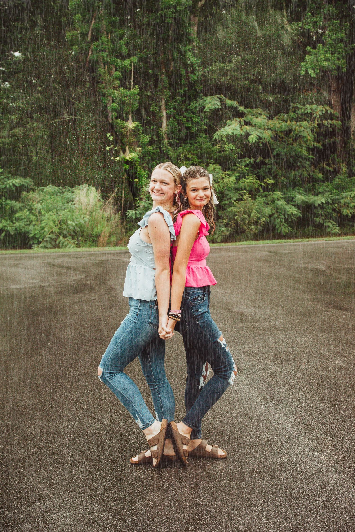 Two friends in casual denim outfits pose back-to-back on a paved path with forest scenery in the background.