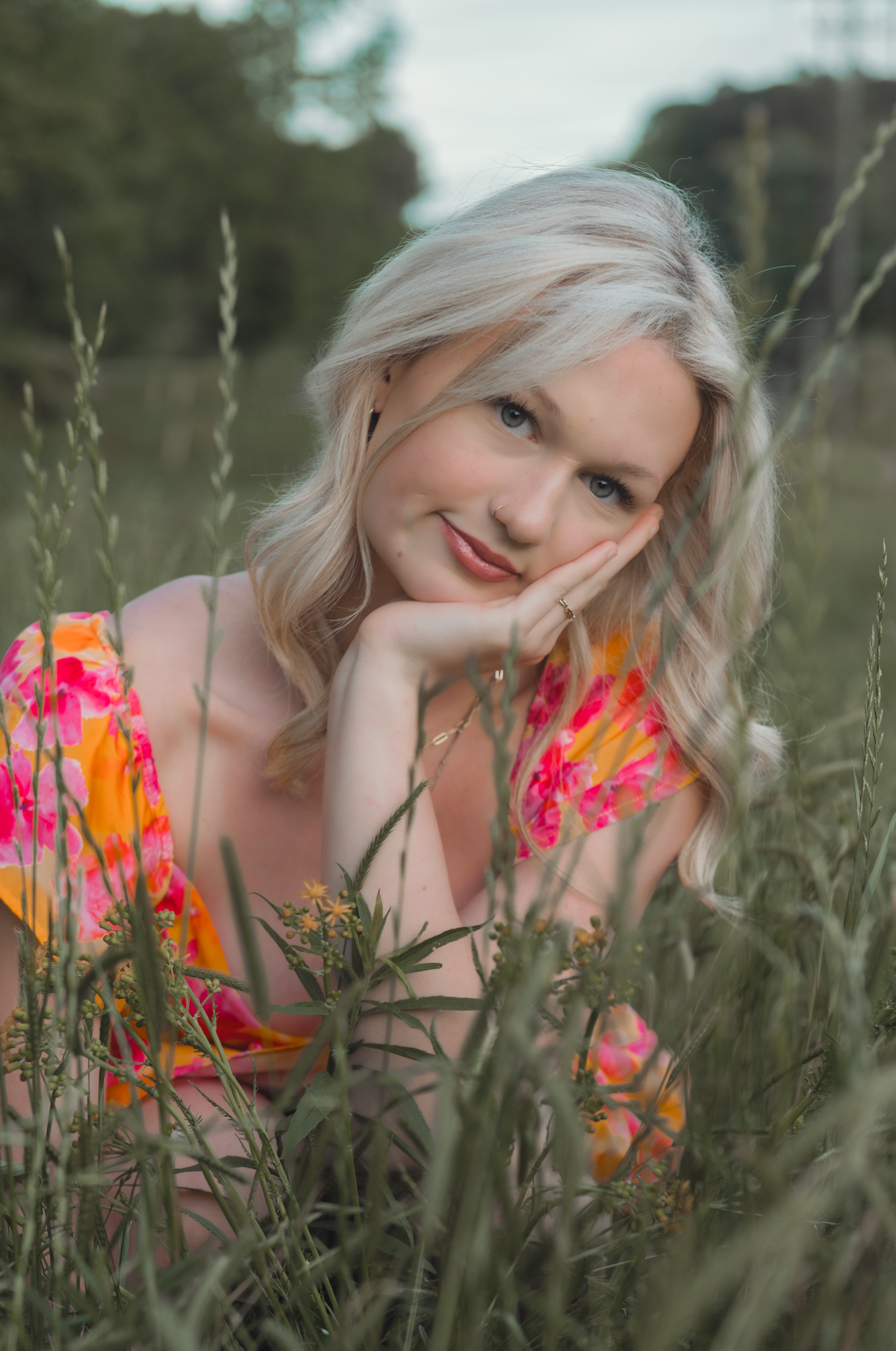 Woman with platinum blonde hair wearing vibrant floral dress poses in tall grass on a sunny afternoon.