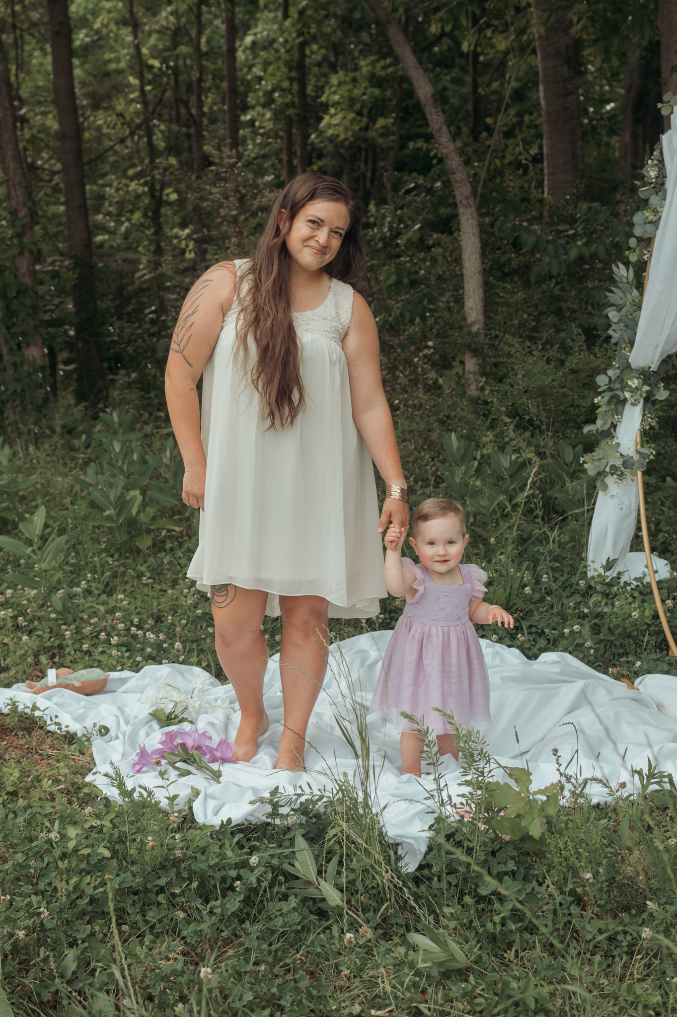 A mother and small child in light dresses stand together on a white blanket in a wooded outdoor setting.