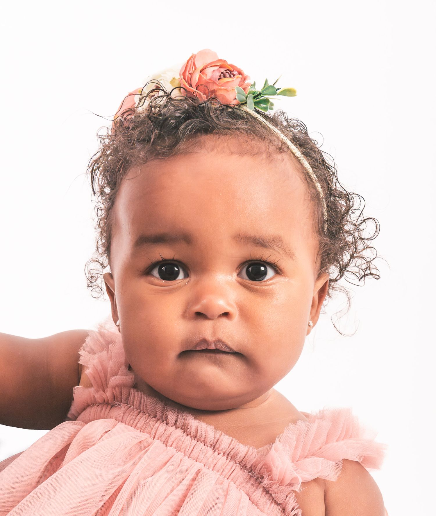 A baby wearing a pink ruffled dress and delicate flower headpiece poses against a white background.