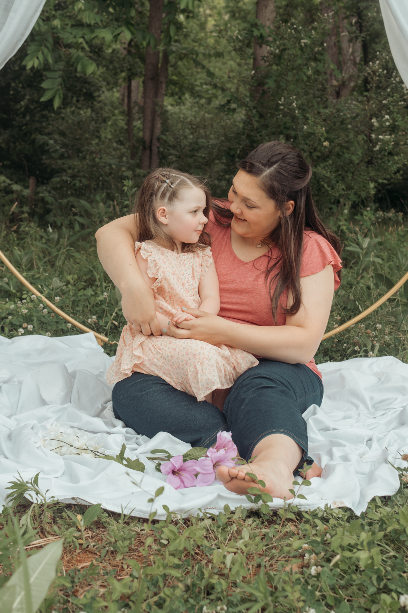 Sweet outdoor portrait of a parent and child sitting together on a white blanket with pink flowers in a wooded setting.
