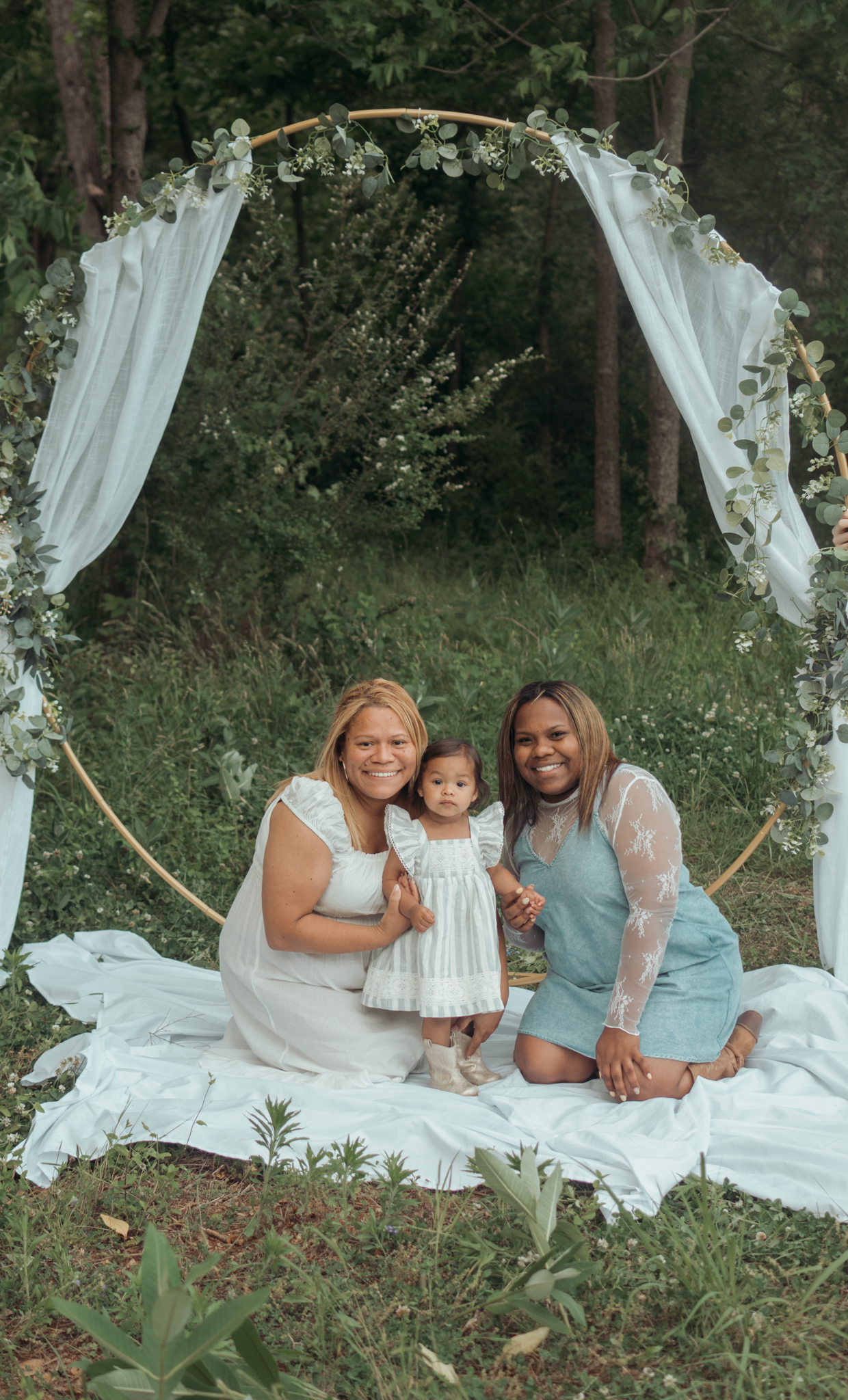 A decorative white arch with flowing fabric and floral accents creates a romantic woodland photo setting on a white blanket.