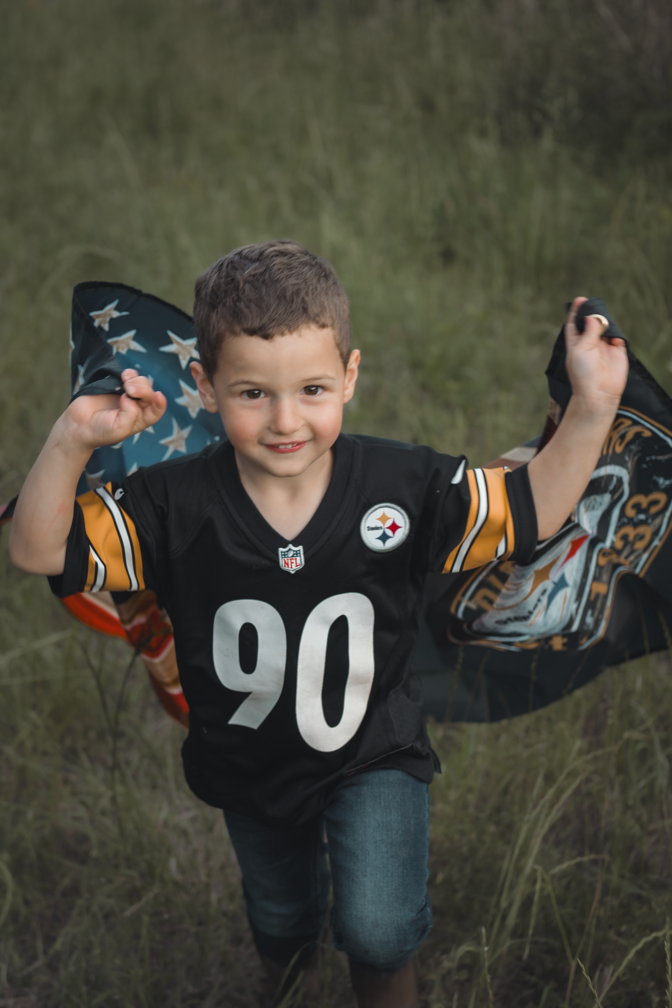 Young football fan wearing Pittsburgh Steelers jersey number 90 holds team flags while standing in a grassy field.