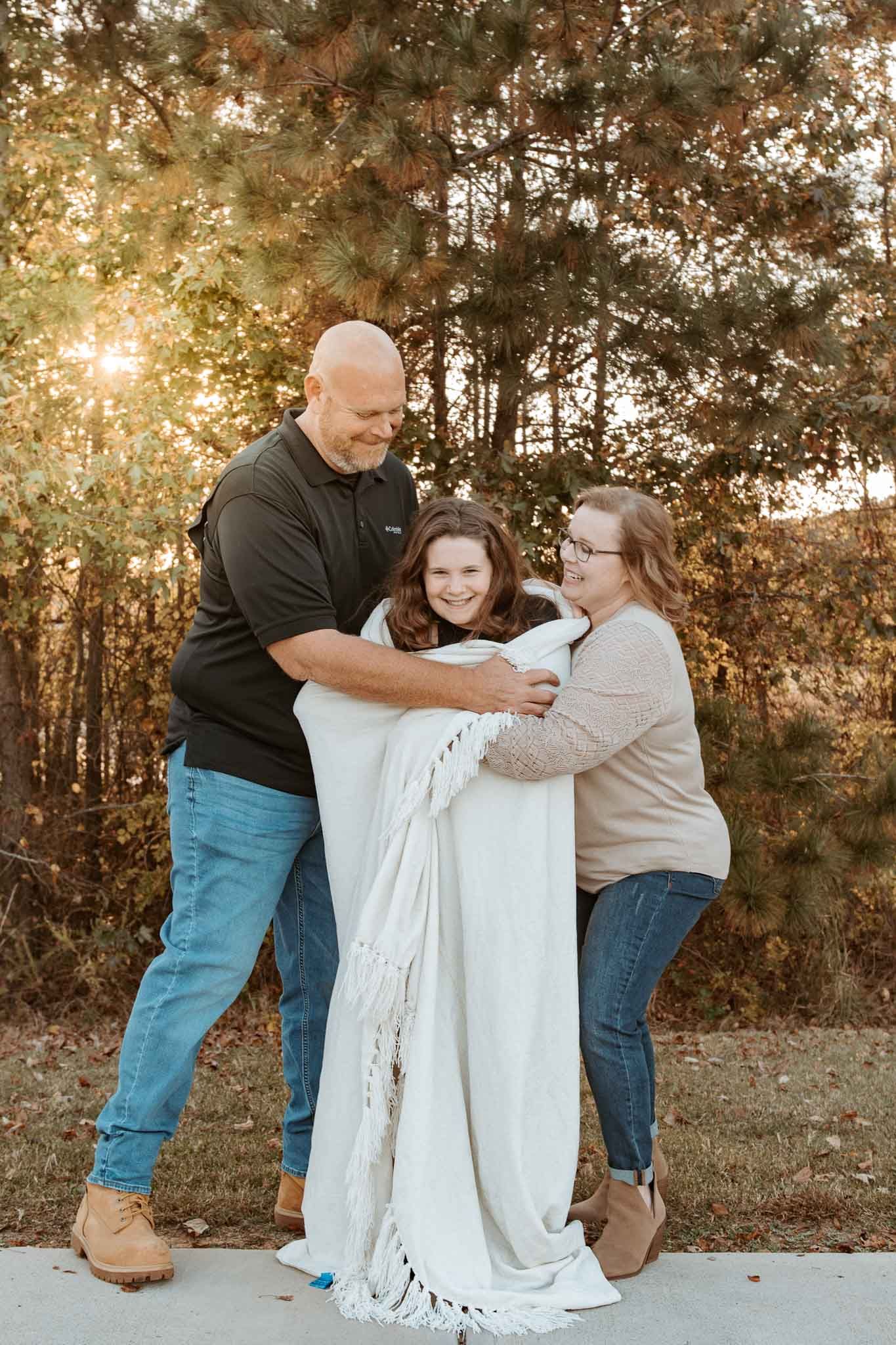 A family shares a warm embrace while wrapped in a white blanket during sunset in a wooded setting.