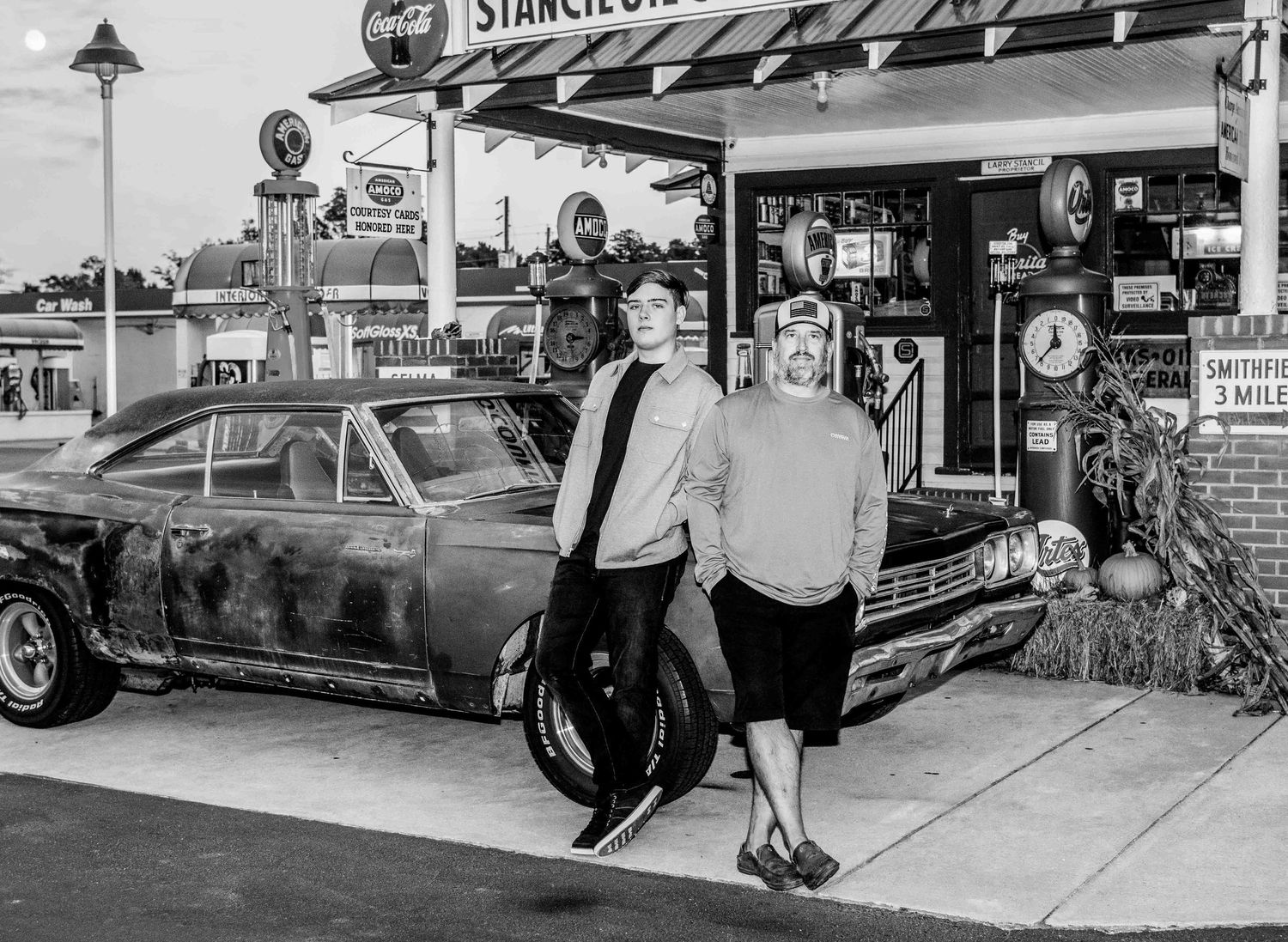 Black and white photo of customers at a vintage Sinclair gas station with classic car and retro signage in the 1950s.