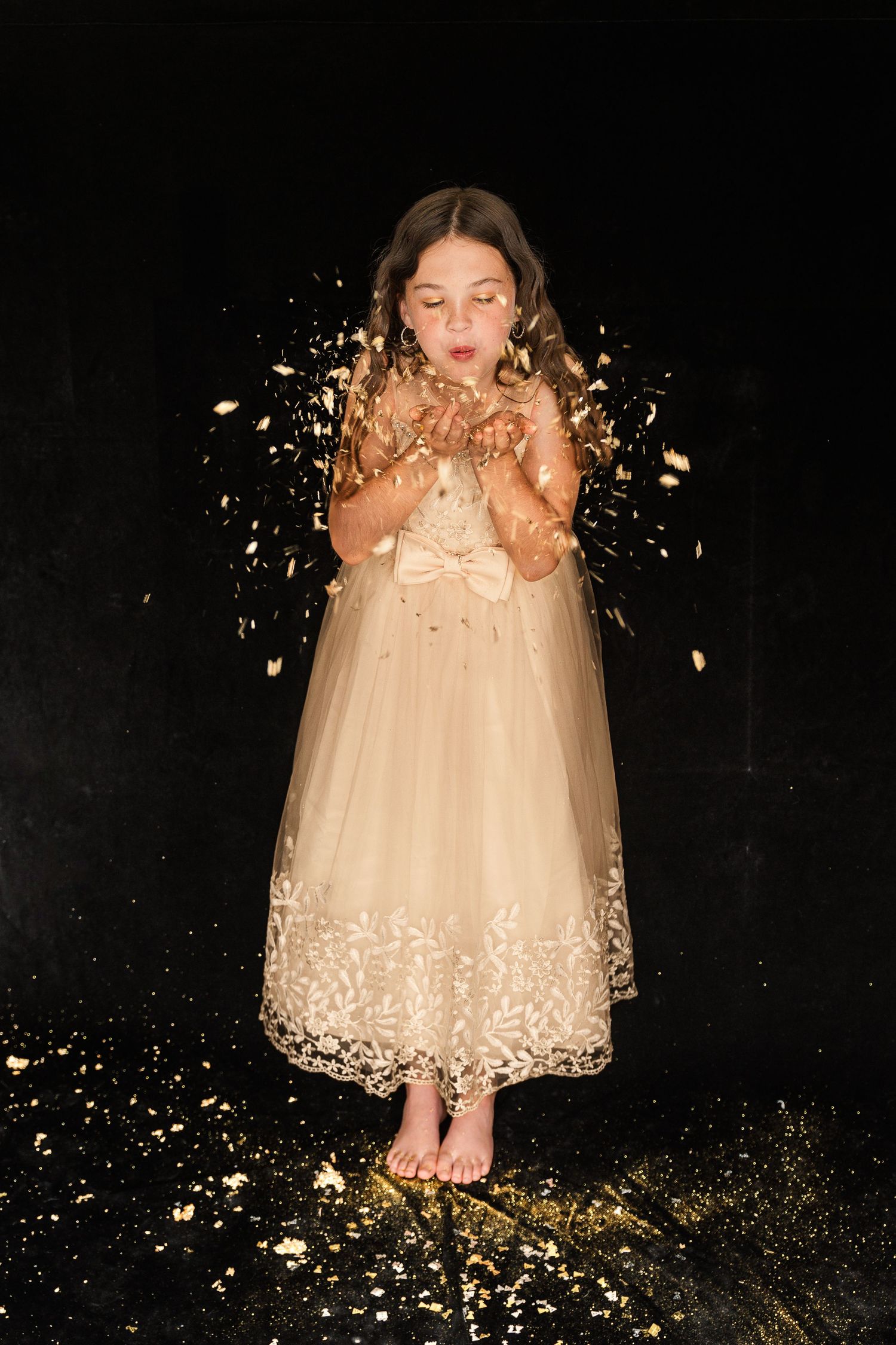 A person in a beige tulle dress stands barefoot while golden glitter sparkles and falls around them against a dark background.