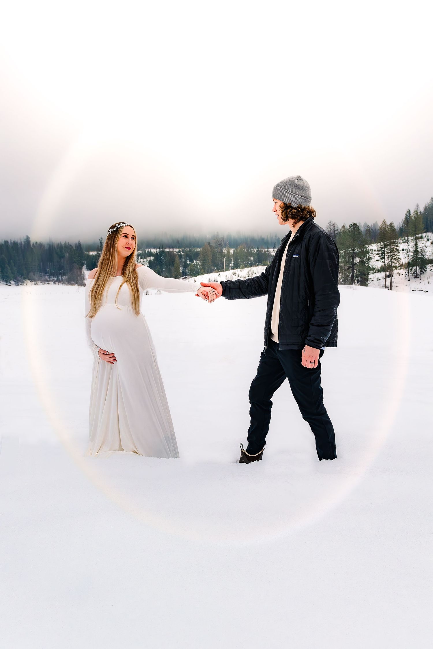 A couple holds hands in a snowy winter landscape with the bride wearing a flowing white dress and groom in dark attire.