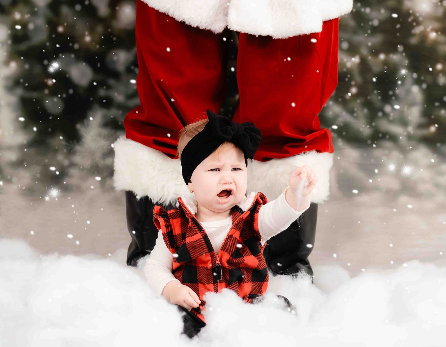 A festive holiday photo of a small child in a red plaid vest sitting in snow next to Santa's legs.