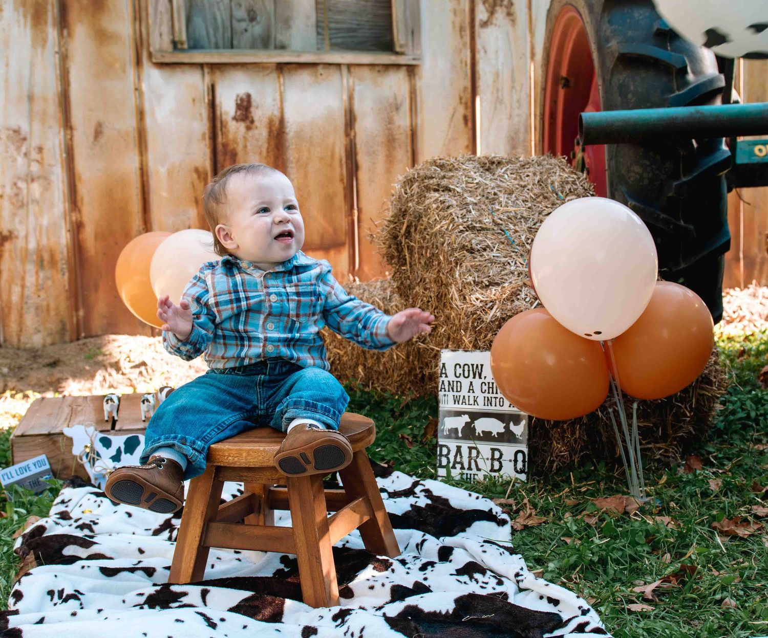Baby in plaid shirt sits on wooden rocking horse with orange and pink balloons against rustic backdrop and hay bale.