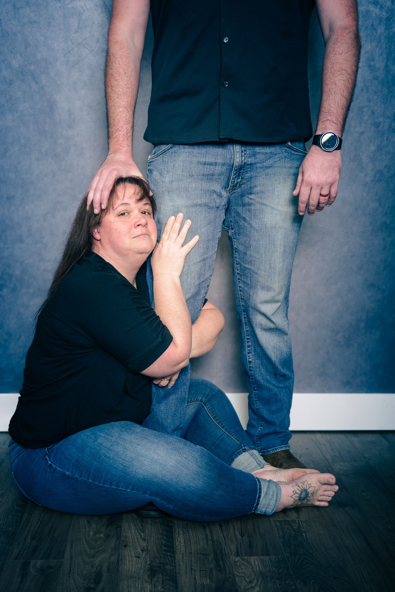 A couple poses casually on a wooden floor with one seated and one standing against a blue wall.