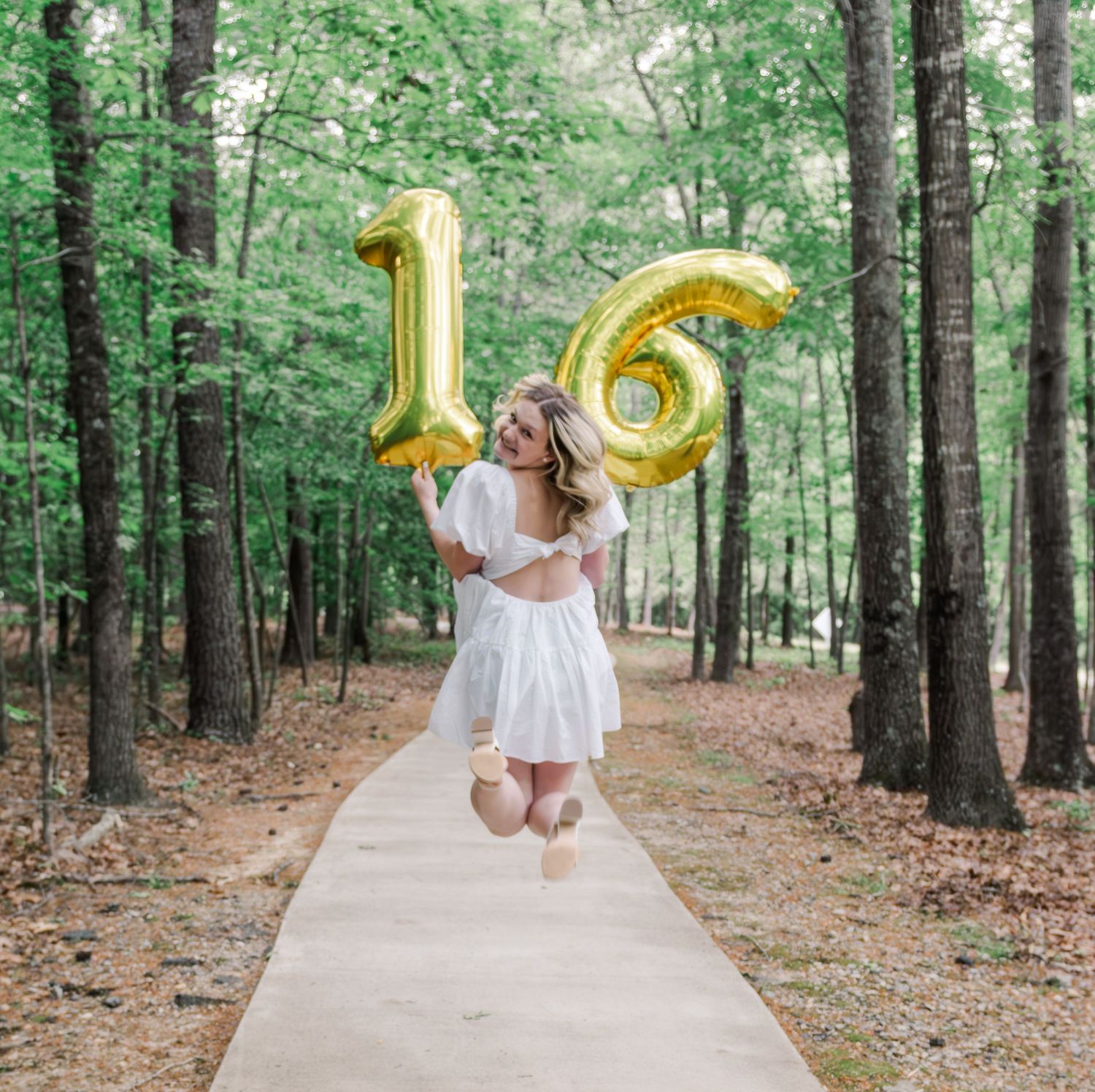 Someone in a white dress jumps joyfully on a forest path holding gold number 16 balloons while celebrating.