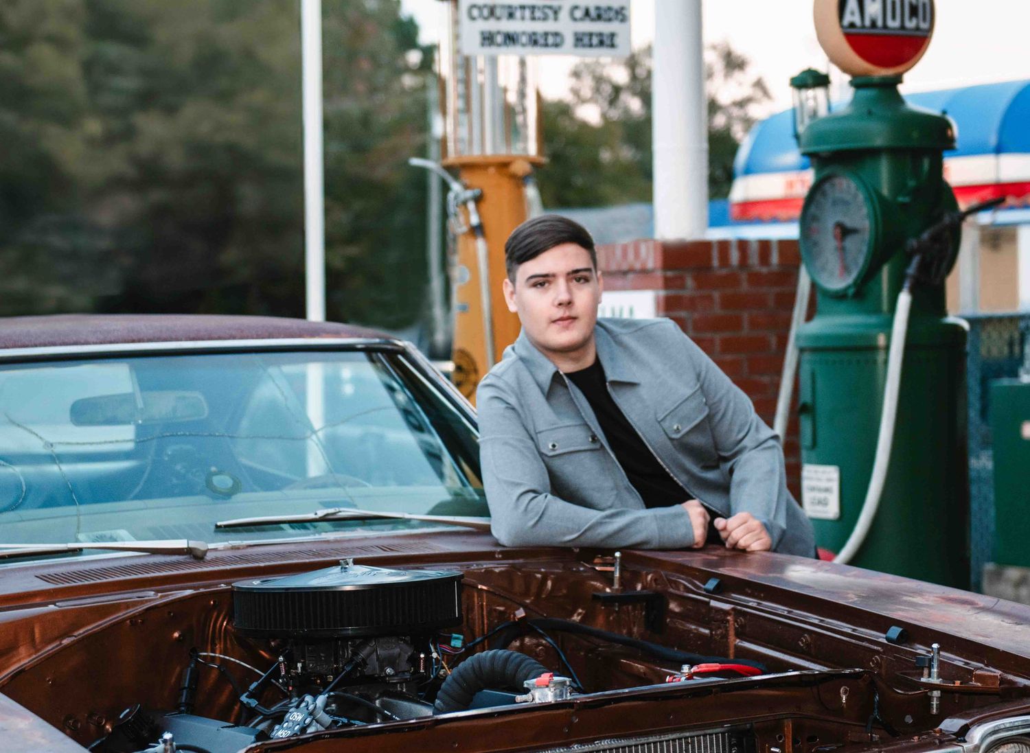 A well dressed figure leans on a vintage car at a retro-styled gas station with antique gas pump in the background.