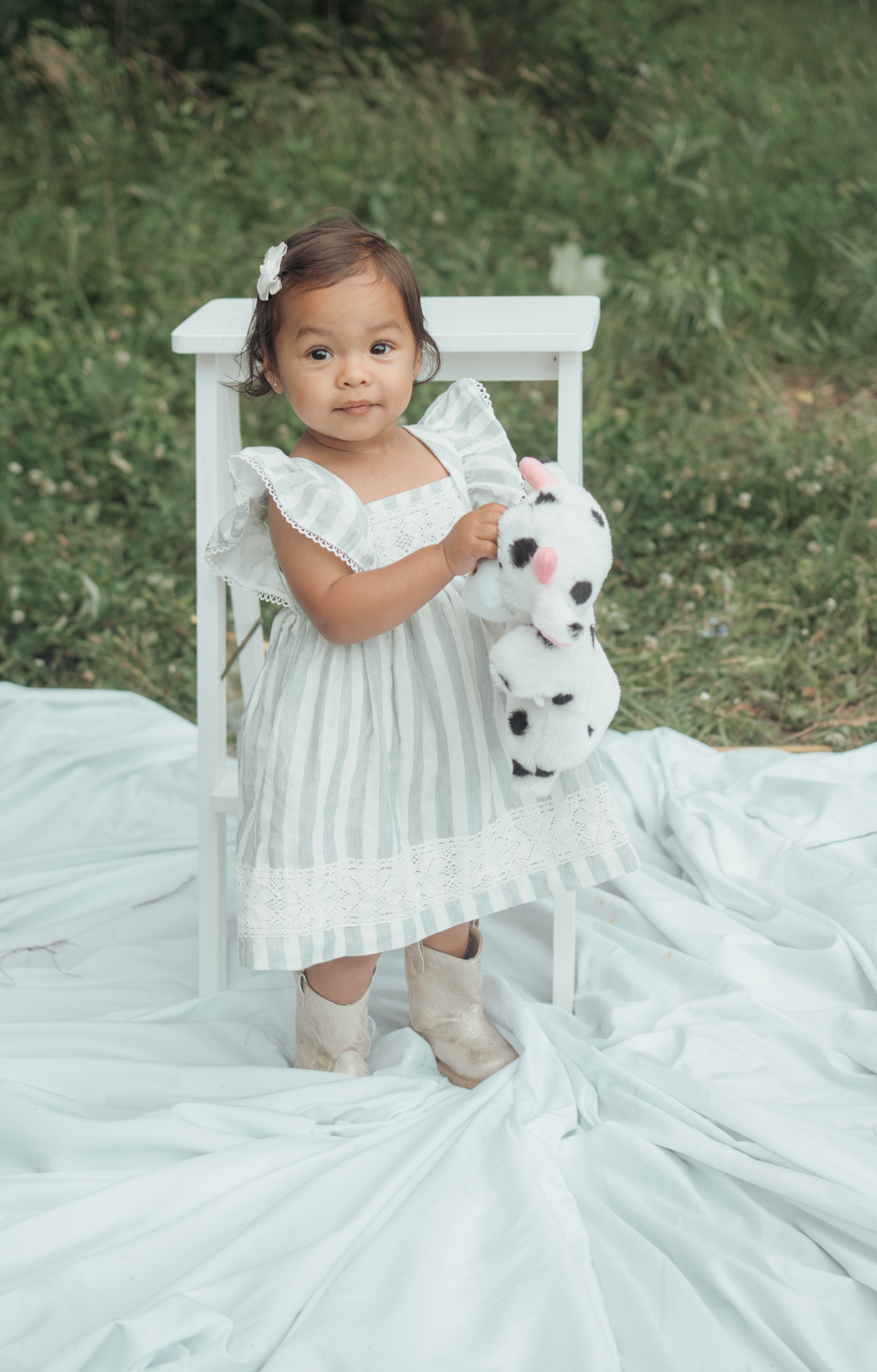 A small child in white dress stands on white fabric outdoors with a plush toy, beside a white wooden chair.