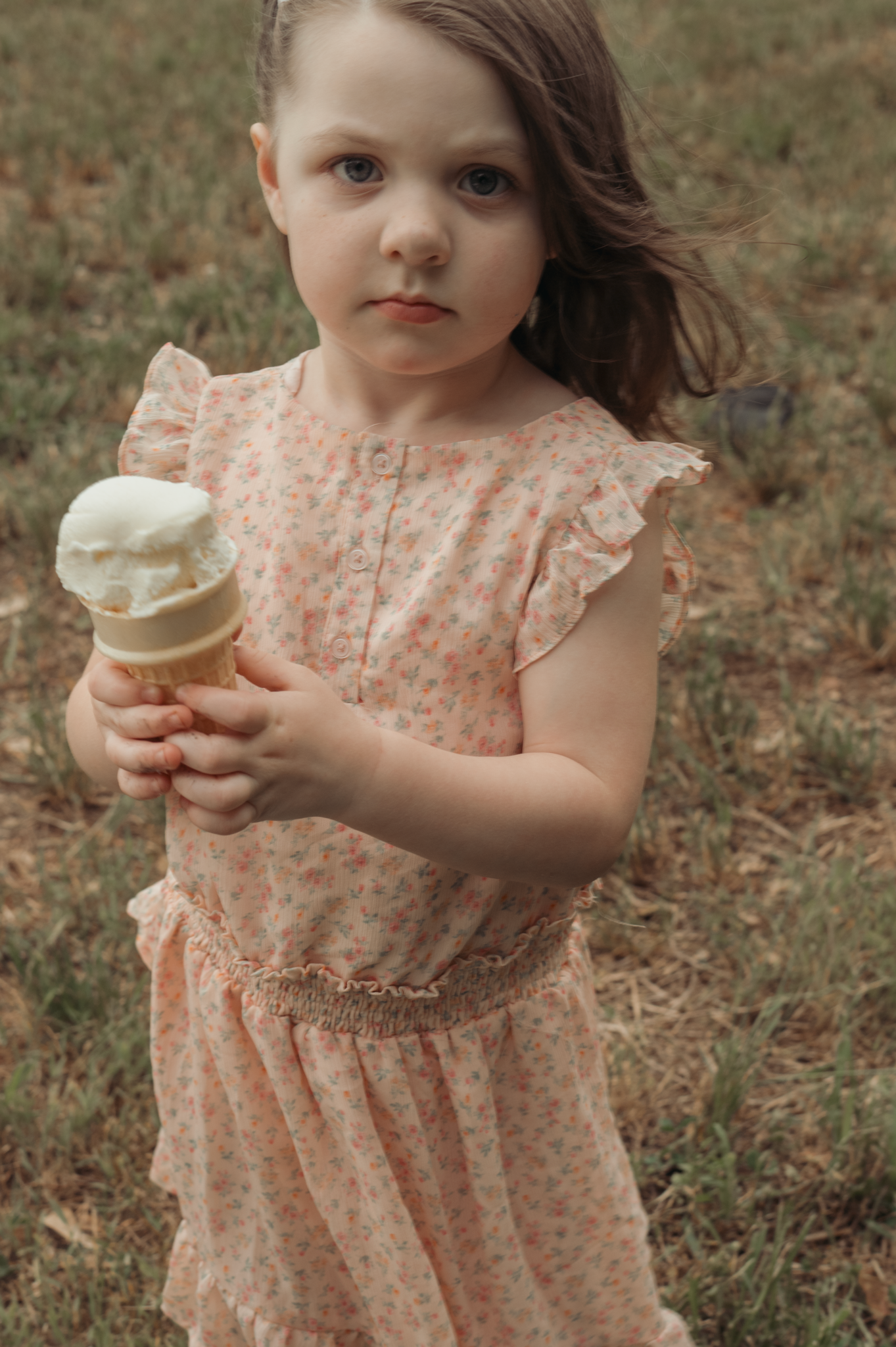 Small child in pink polka dot dress holds vanilla ice cream cone while standing outside on grass.
