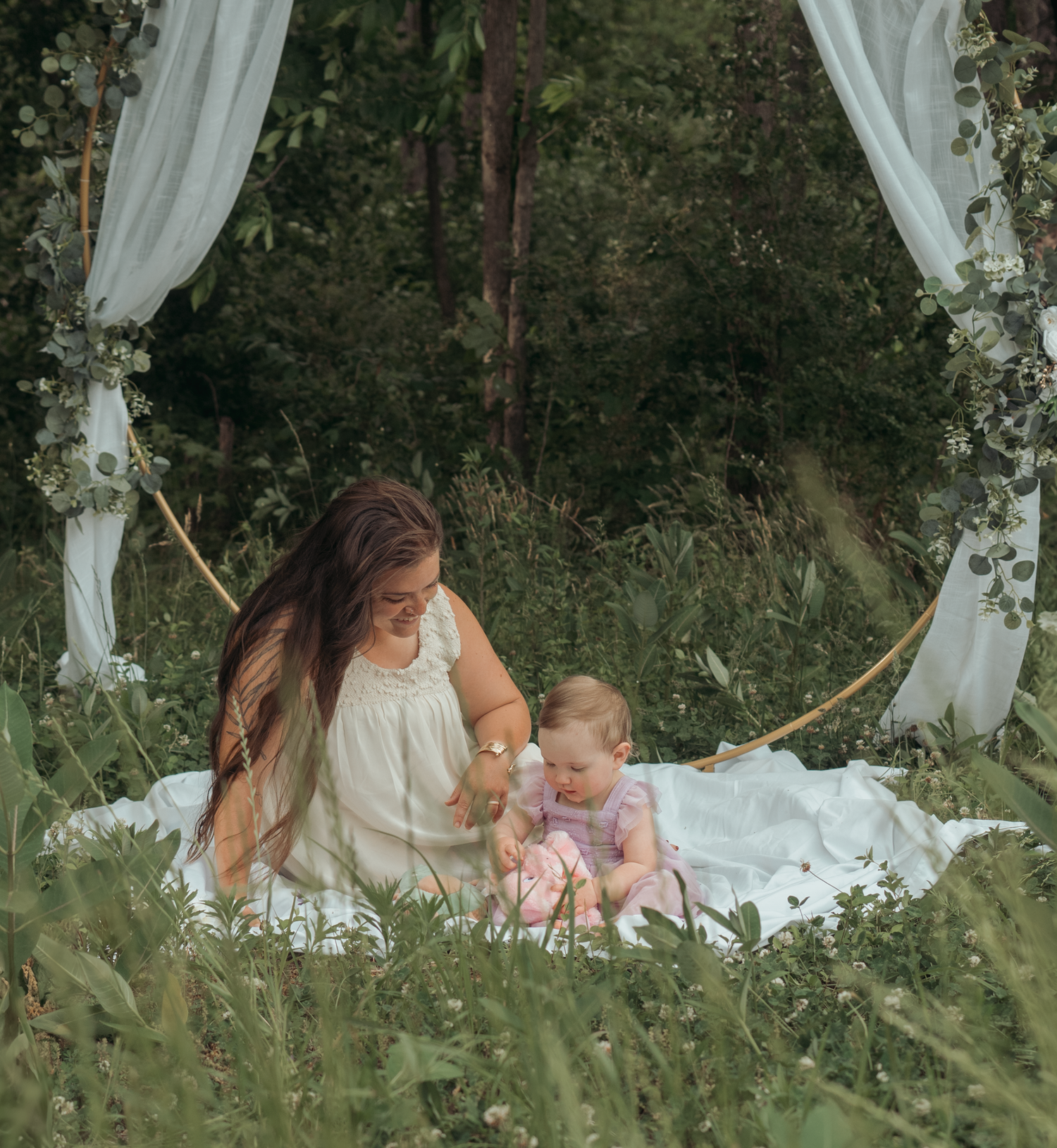 An outdoor portrait under a white fabric arch shows a sweet moment on a blanket in tall grass.
