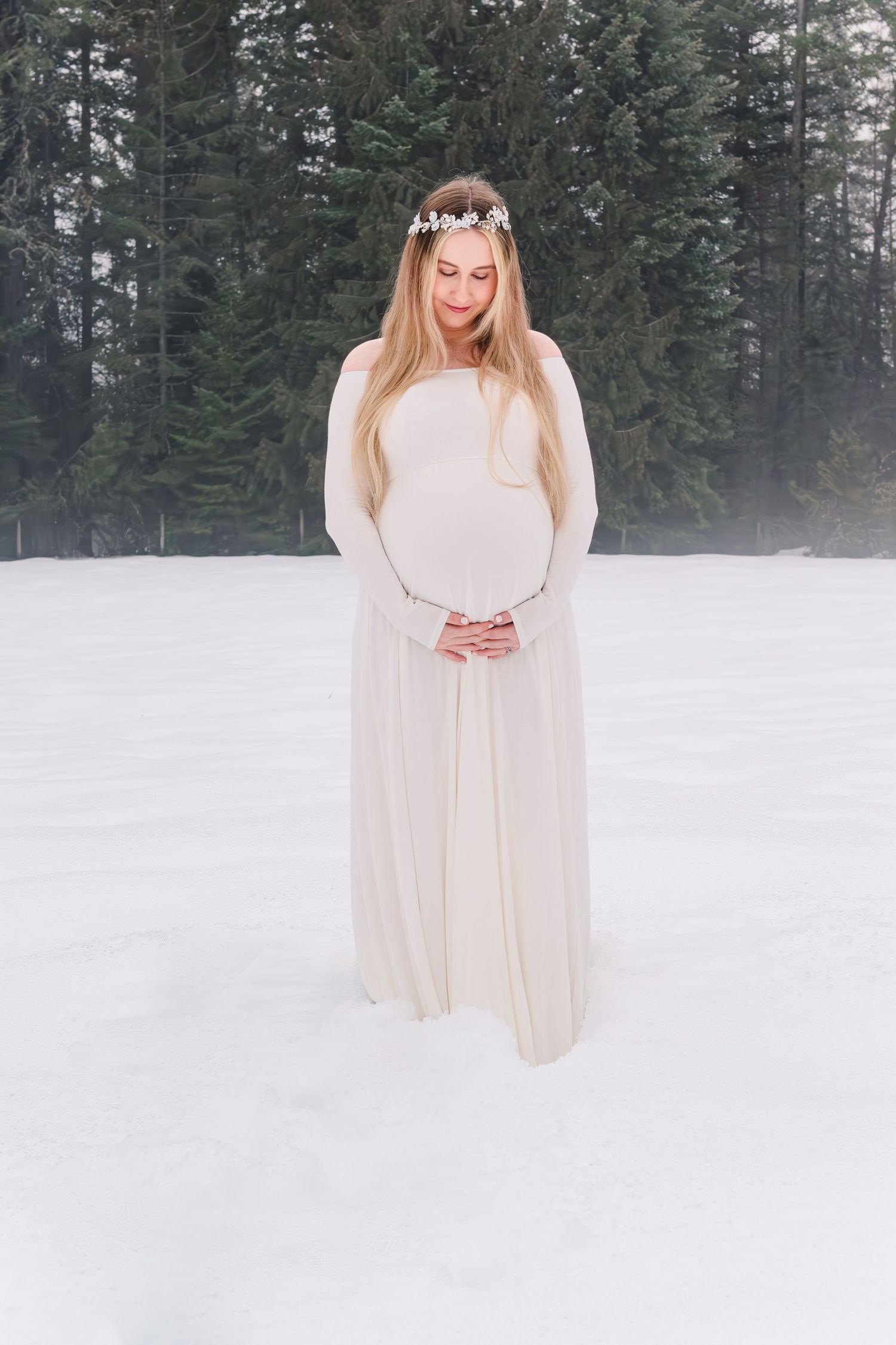 Ethereal winter maternity photograph featuring a flowing white dress and floral crown against a snowy forest backdrop.