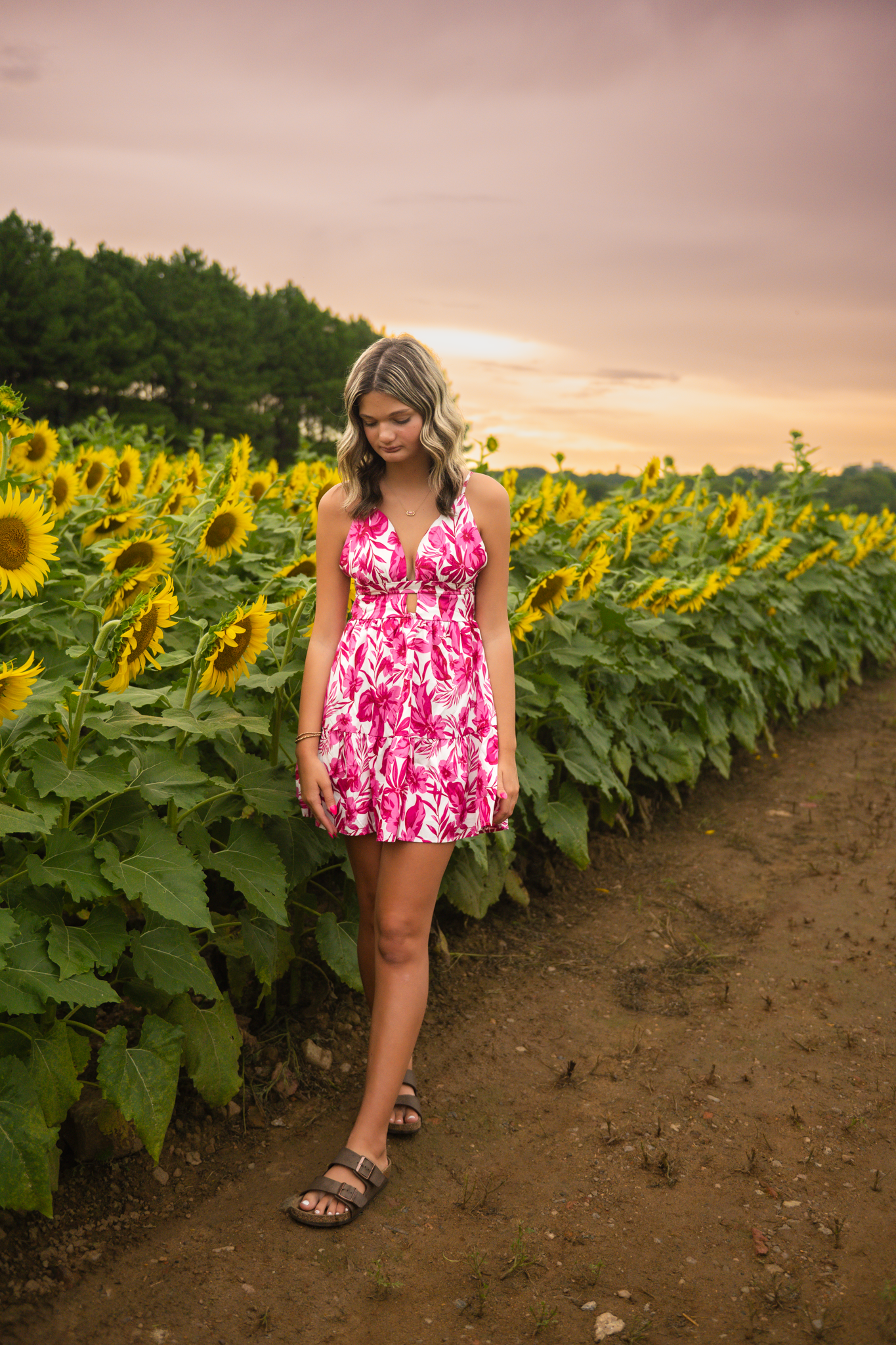 A person in a pink floral sundress standing on a dirt path between rows of tall sunflowers at sunset.