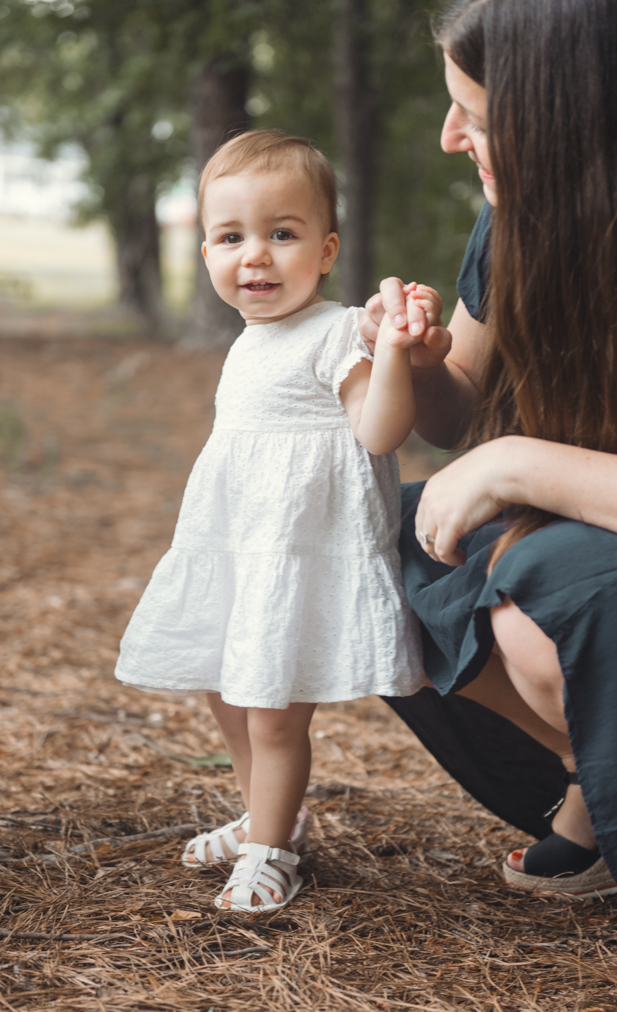 A tender moment between parent and child outdoors in a wooded area, with the little one wearing a white dress.