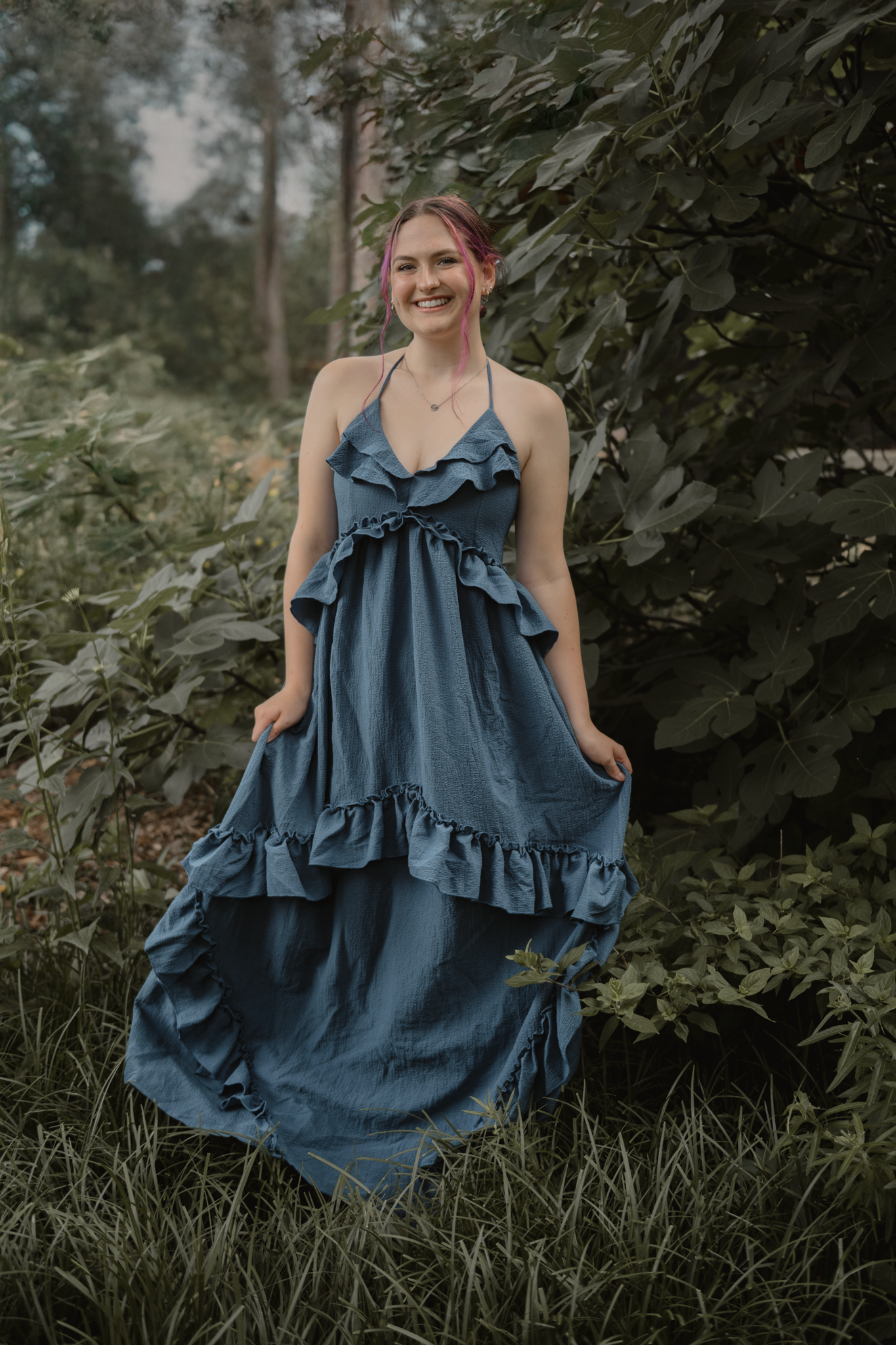 A flowing blue ruffled maxi dress photographed outdoors in a natural woodland setting with soft lighting.