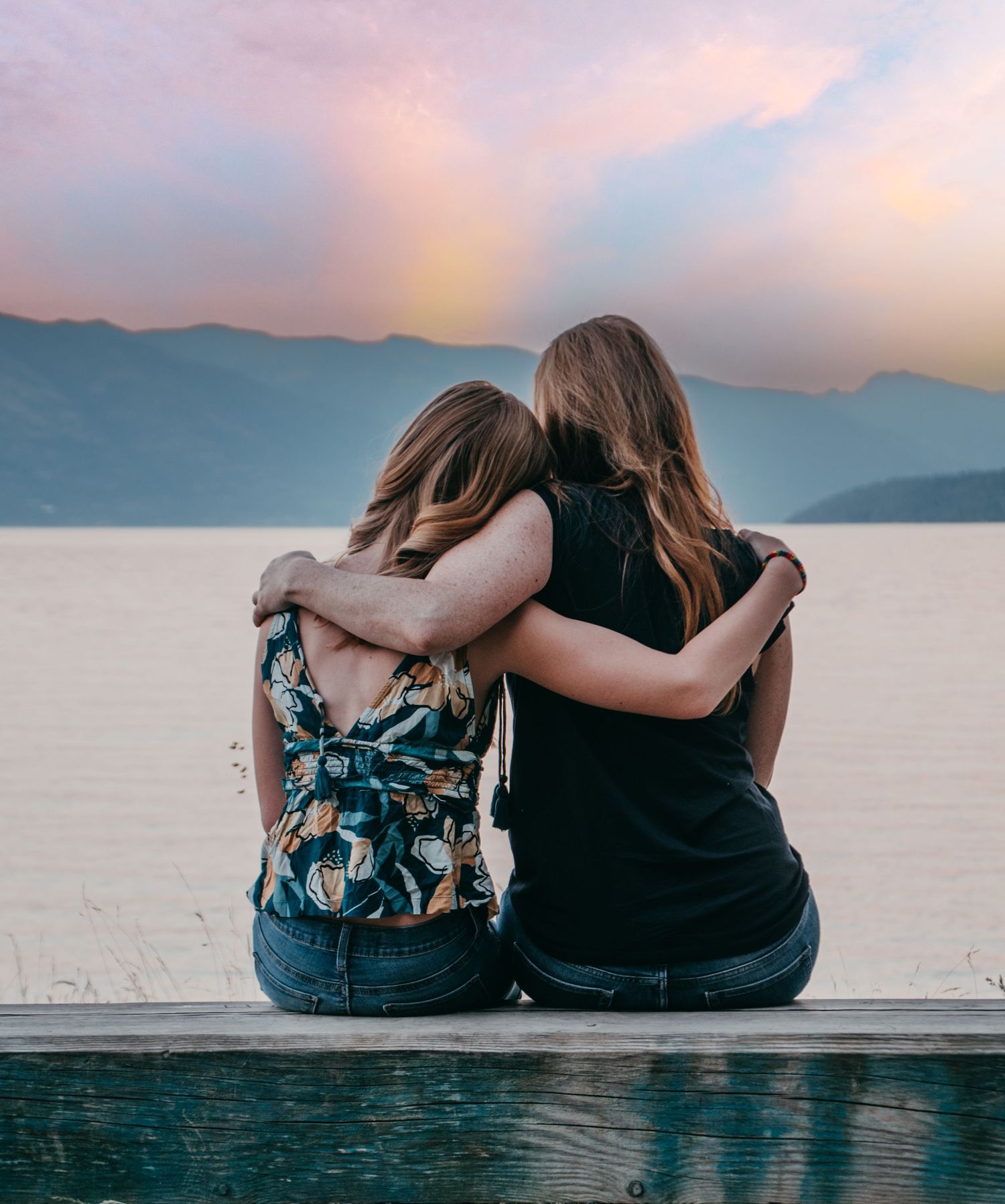 Two people sit together on a dock overlooking a serene lake with mountains in the background during sunset.