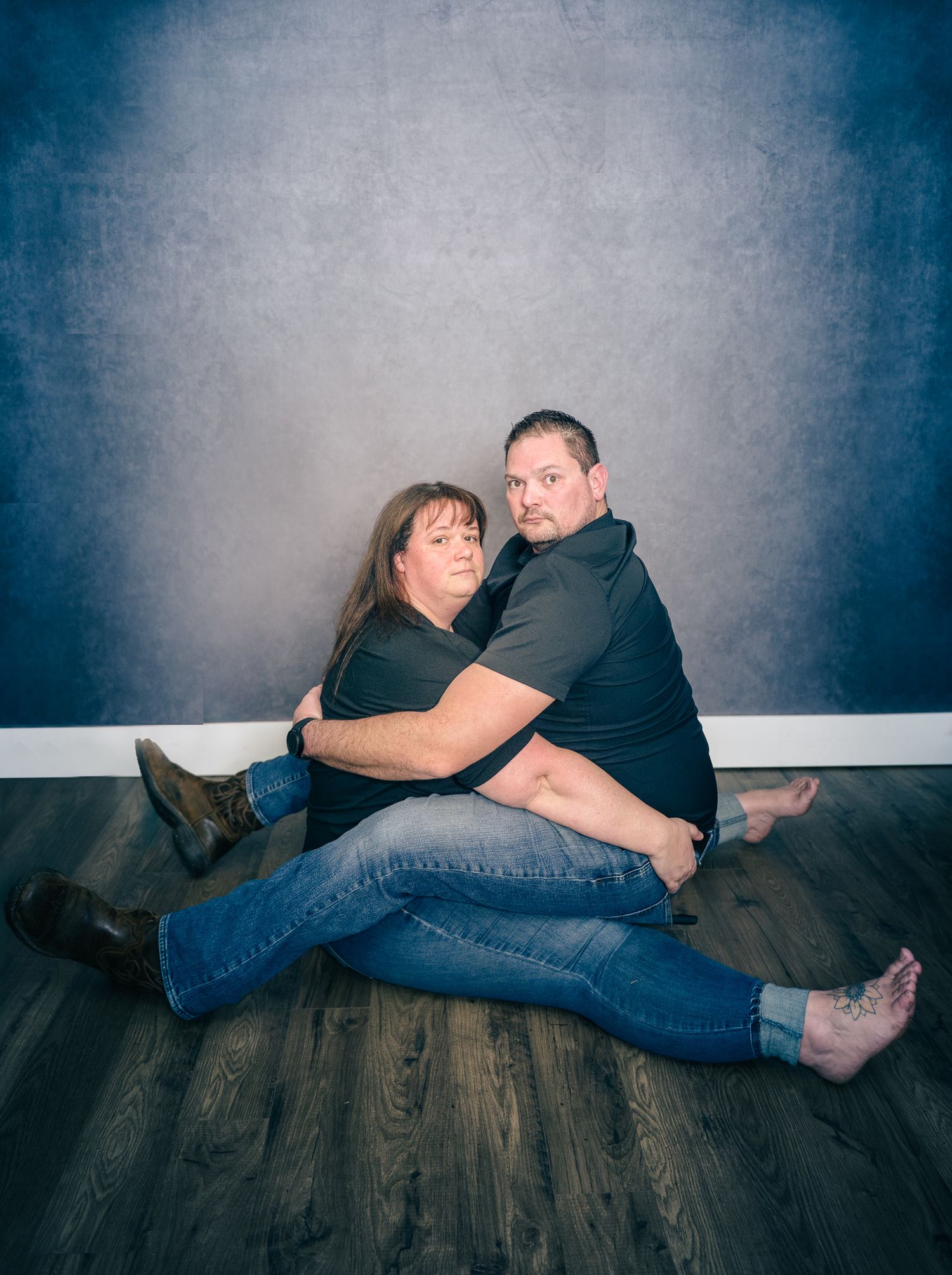A couple in black shirts and jeans pose playfully on a hardwood floor against a blue-gray backdrop.