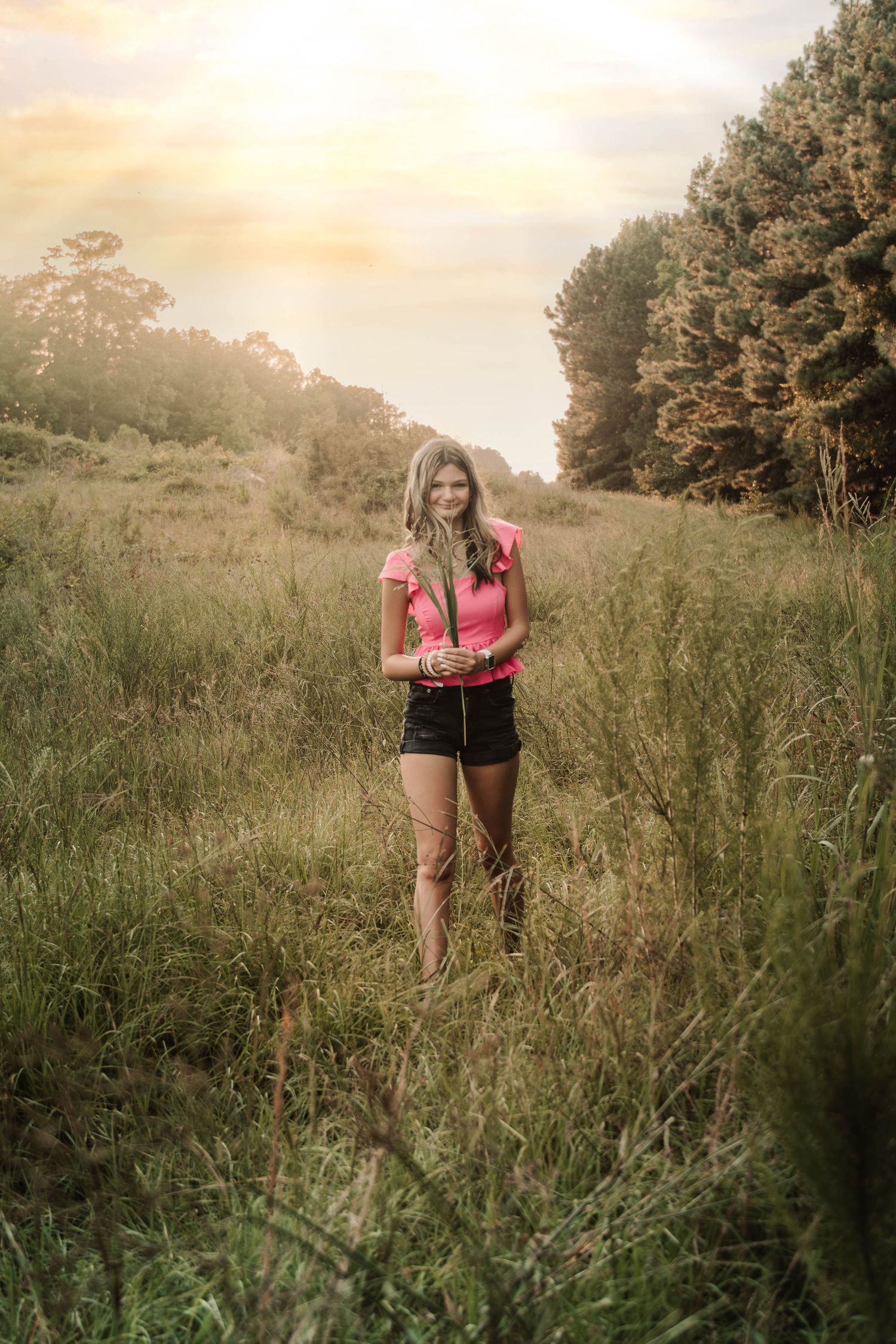 Someone in a pink top and dark shorts stands in tall grass at sunset while golden light illuminates the meadow.