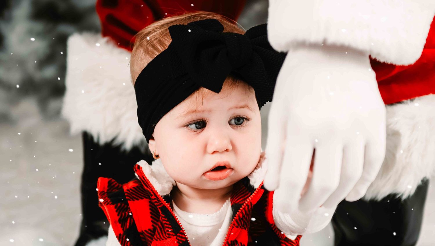A small child in buffalo plaid and a black cap sits in snow next to Santa's red suit during a winter photo shoot.