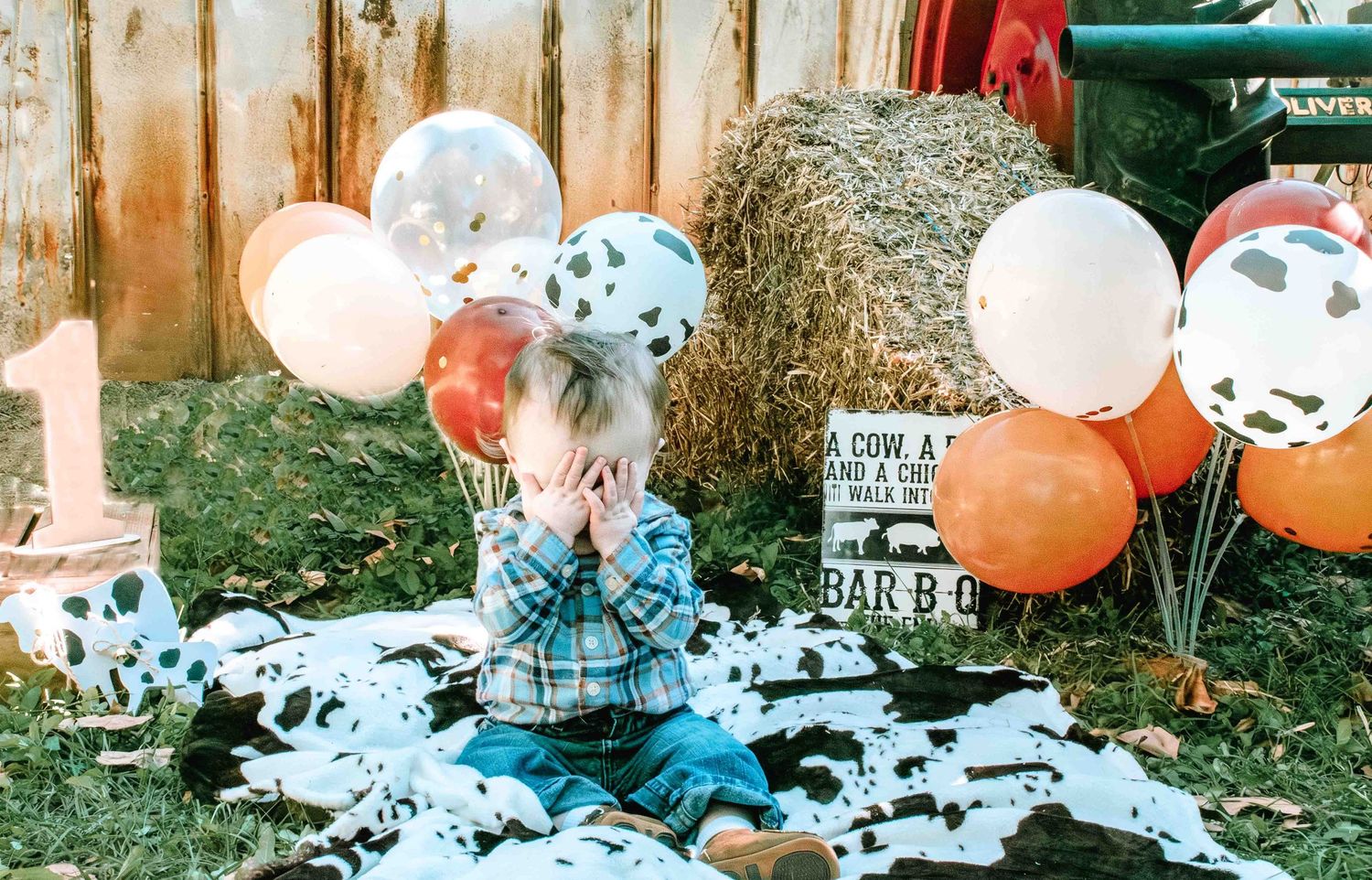 Decorated outdoor party setup with cow print balloons, orange balloons, and hay bales against a rustic wooden fence.