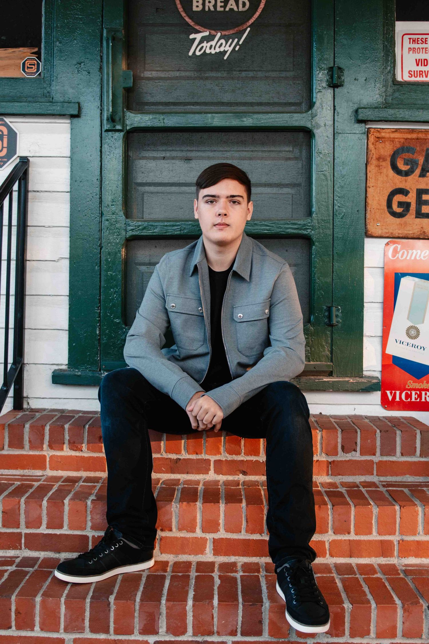 Someone sits on a brick ledge wearing a gray jacket and black pants in front of a vintage storefront with retro signage.