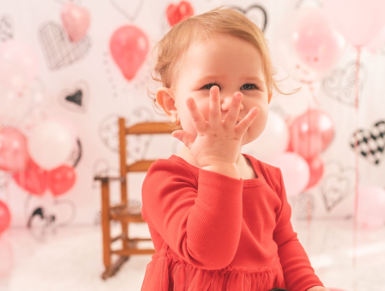 Small child in red dress covering mouth with hand against pink heart decorated background.