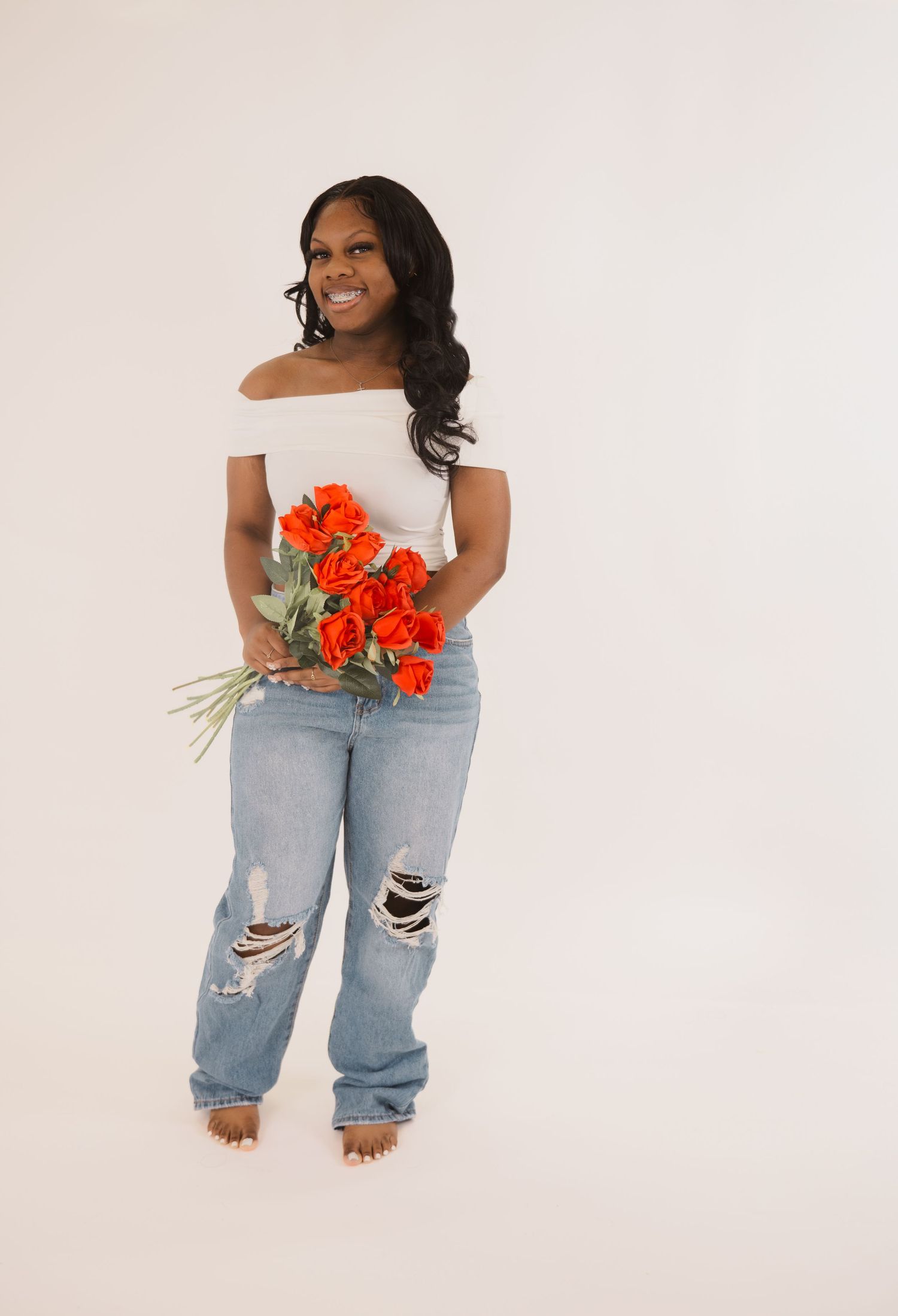 Someone wearing a white off-shoulder top and distressed jeans holds vibrant orange flowers against a light backdrop.