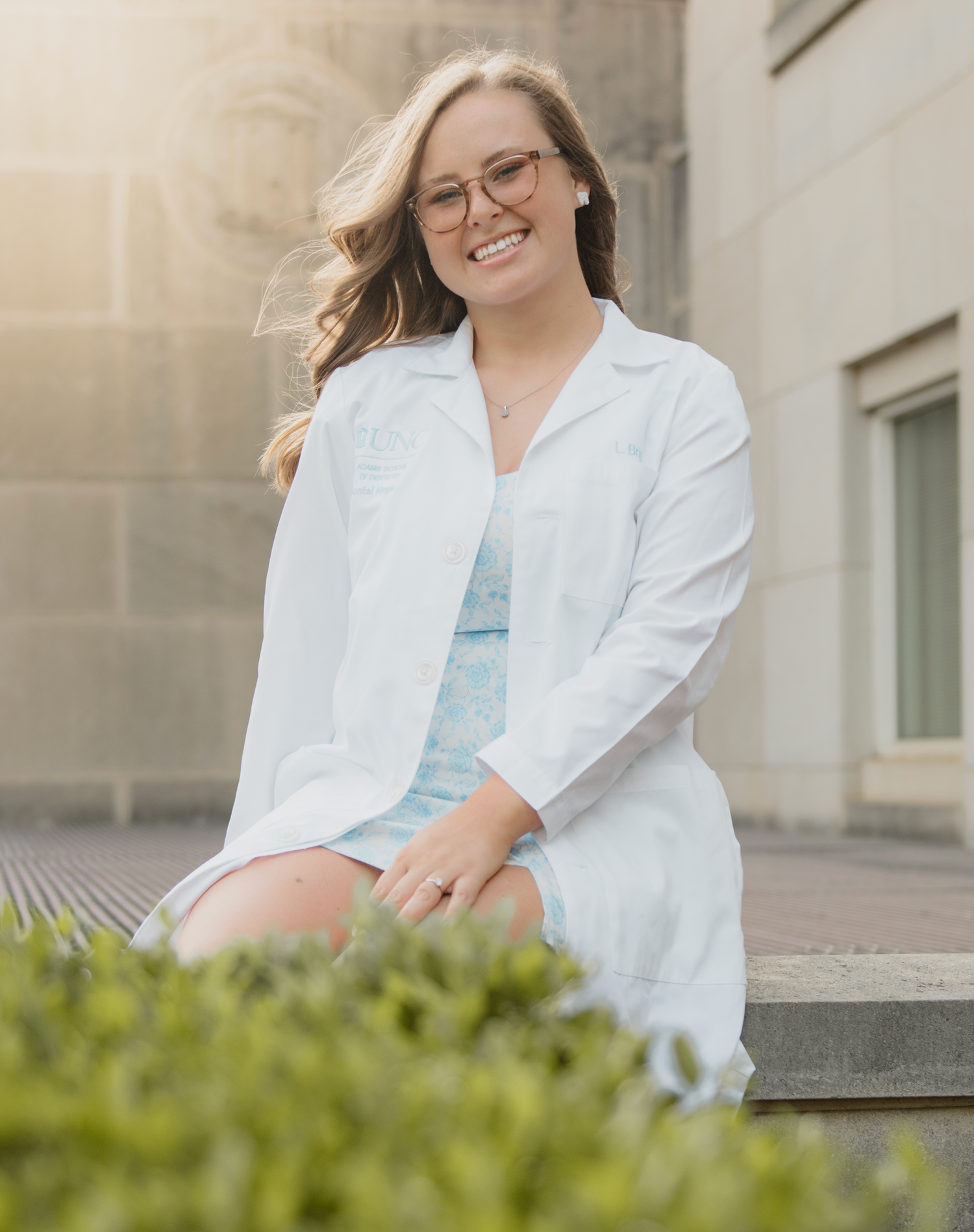 Healthcare professional in a white lab coat sitting outdoors on a sunny day by a building with greenery.