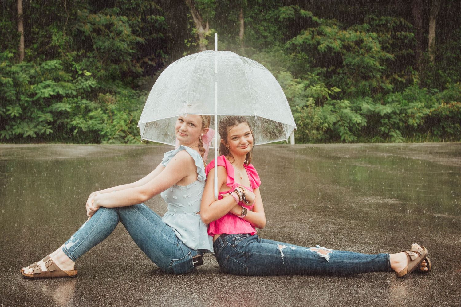 Two friends sit back-to-back under a clear umbrella on wet pavement while enjoying a rainy day outdoors.
