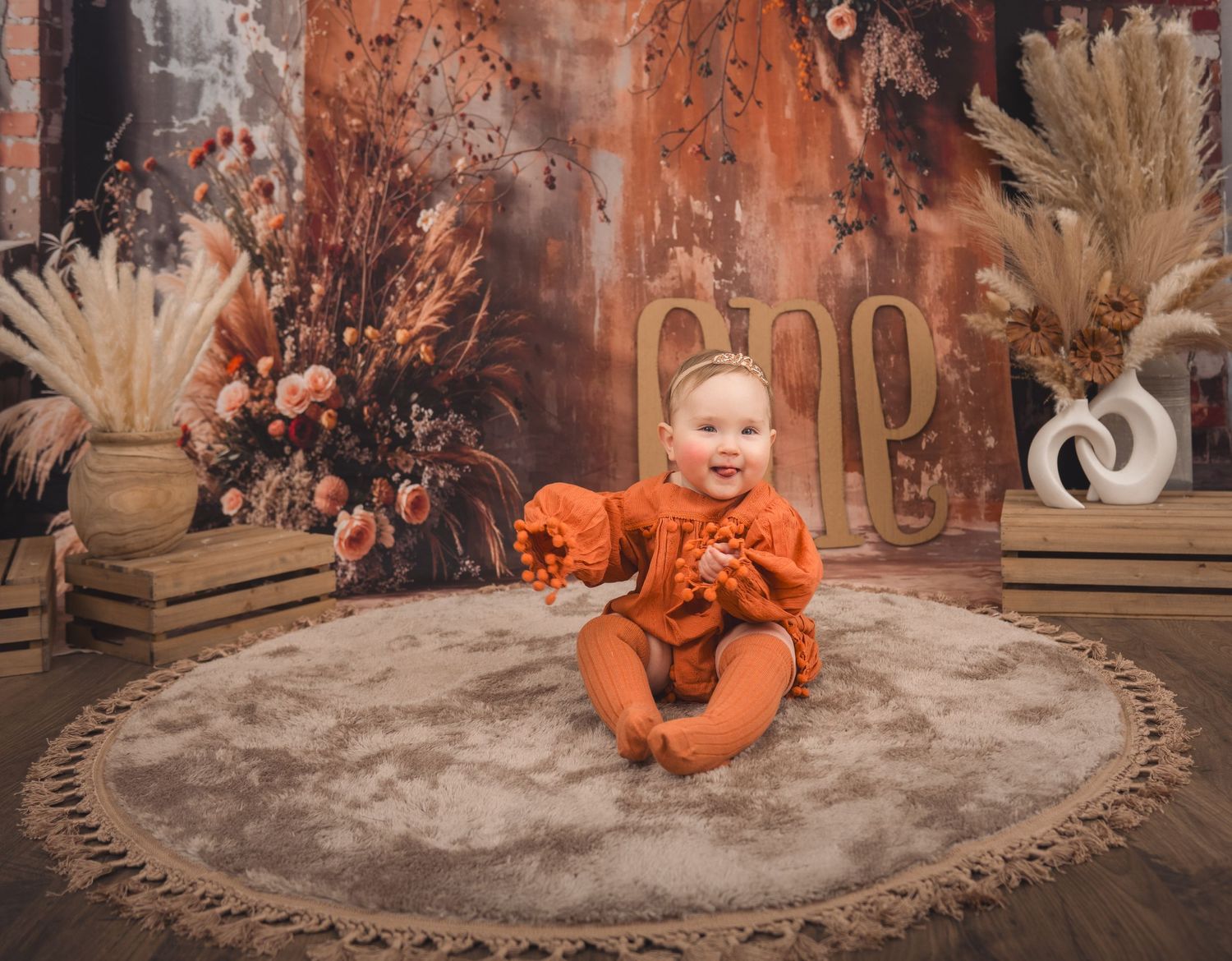 Small child in bright orange outfit sits on round rug amid autumn-themed decor with dried flowers and wooden accents.