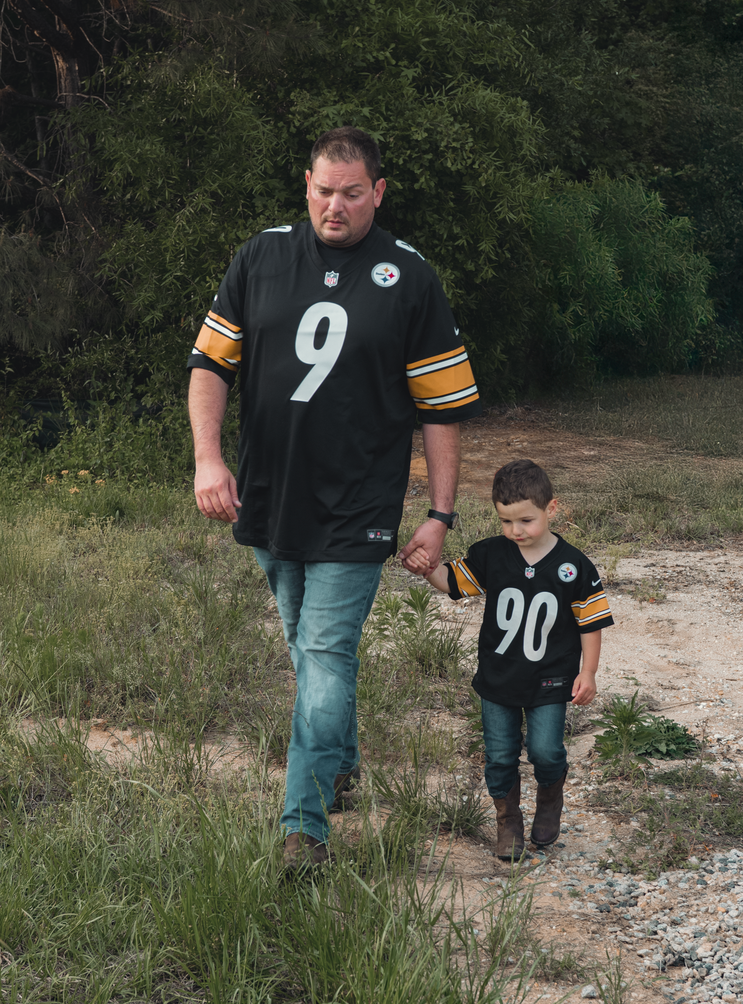 Two Pittsburgh Steelers fans walk together on a dirt path wearing matching jerseys numbered 9 and 90.