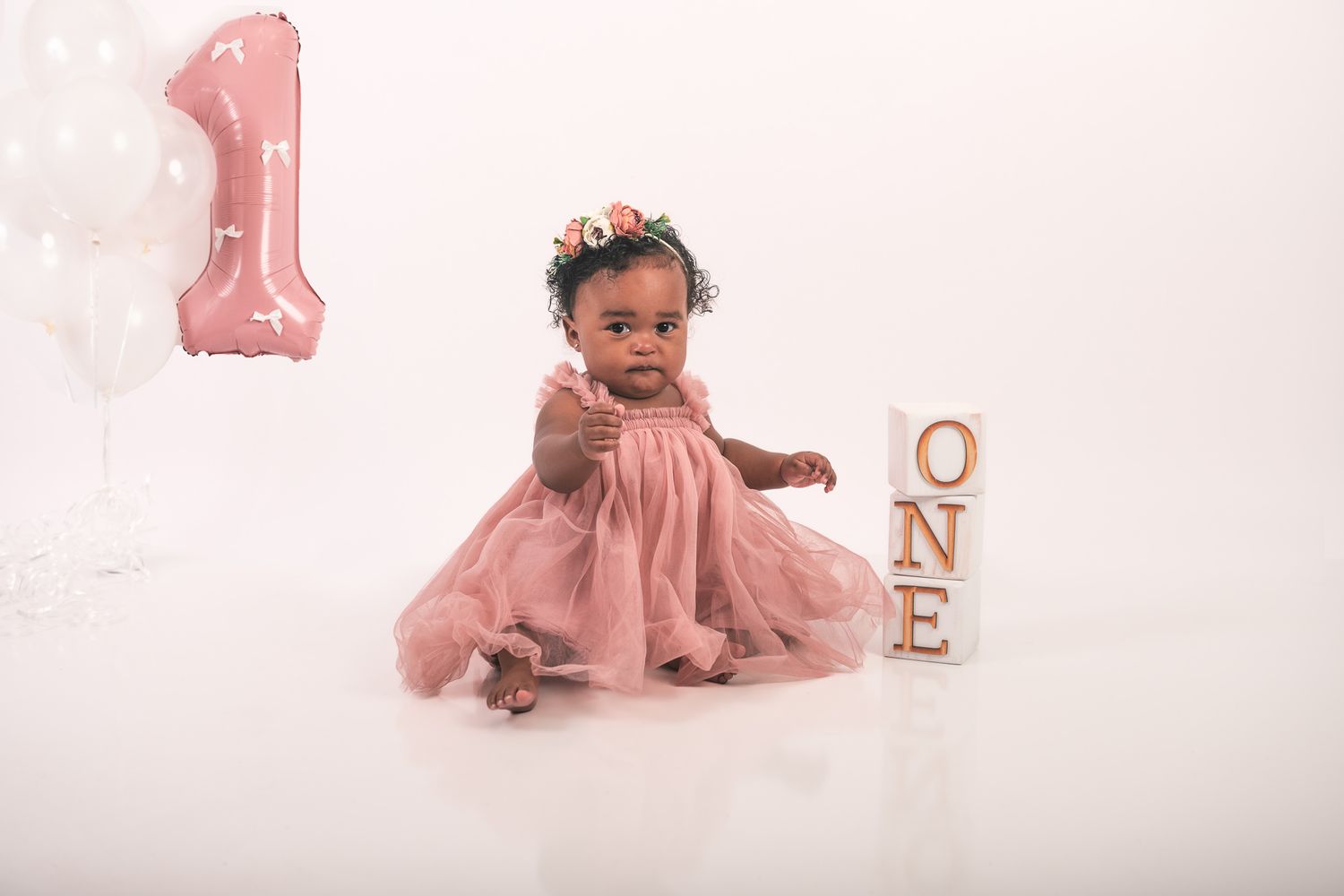 Baby wearing a dusty pink dress sits on white backdrop near rose gold 'ONE' letters and number 1 balloon decoration.