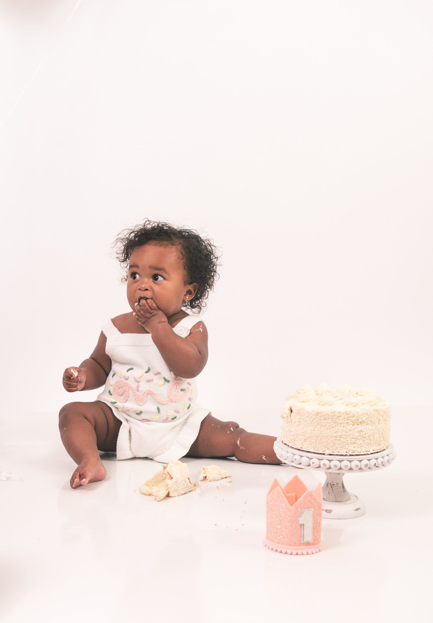 A baby wearing a white onesie sits on the floor with a white cake and pink decorations for a first birthday celebration.