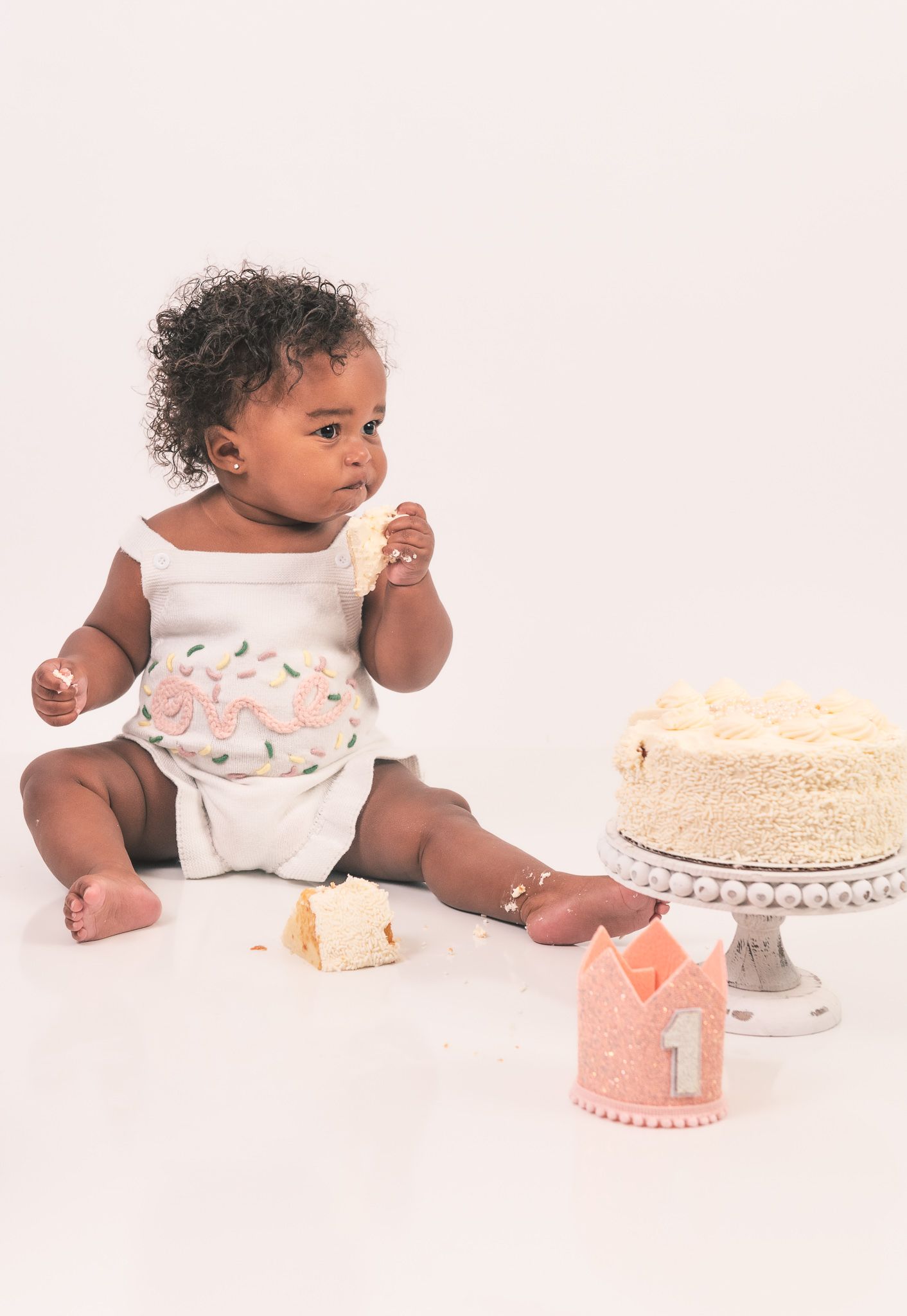 Baby in white floral dress sitting beside white birthday cake and pink crown on light pink background.