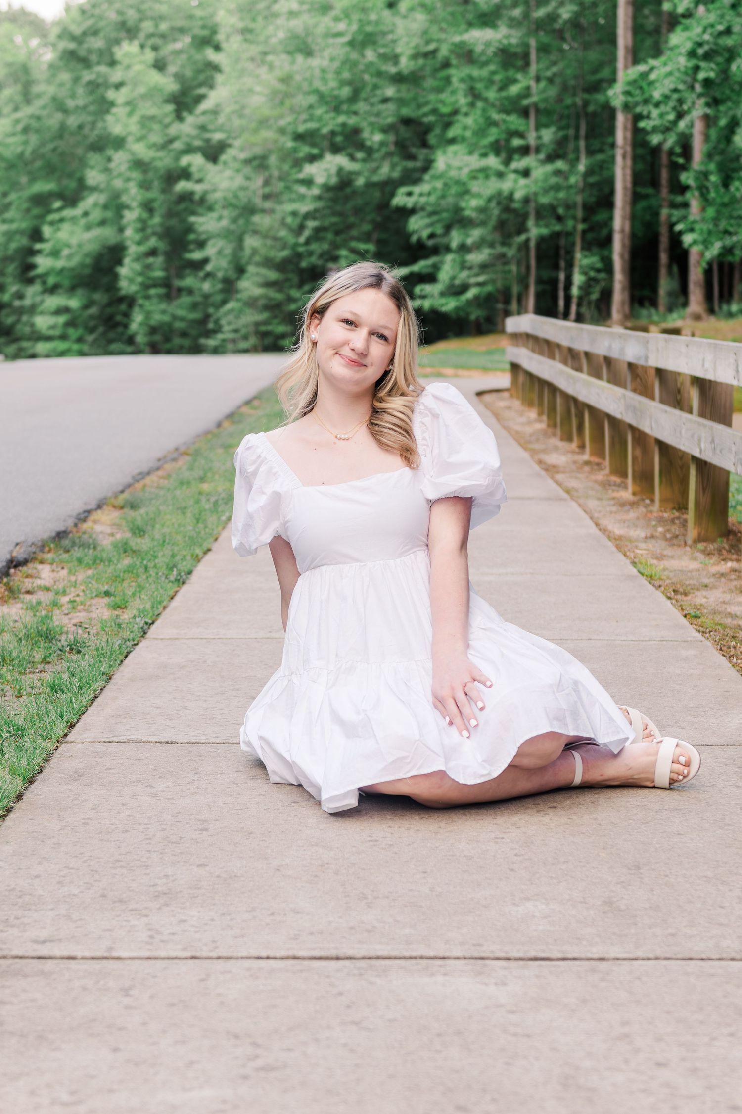 A romantic outdoor portrait of someone in a white puffy-sleeved dress sitting on a concrete path near a wooden railing.