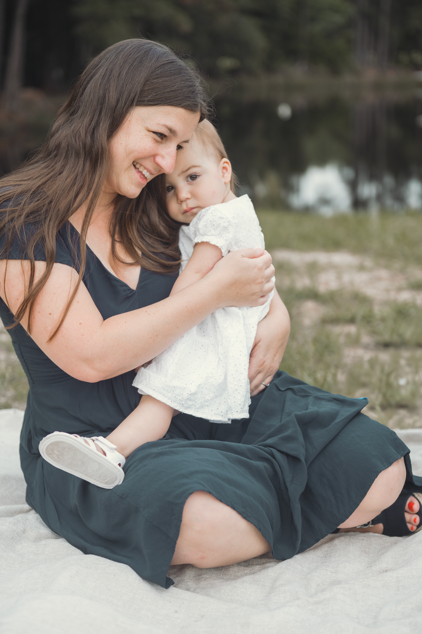 A parent wearing a navy dress sits on a blanket outdoors while holding a baby in a white outfit against a lake backdrop.