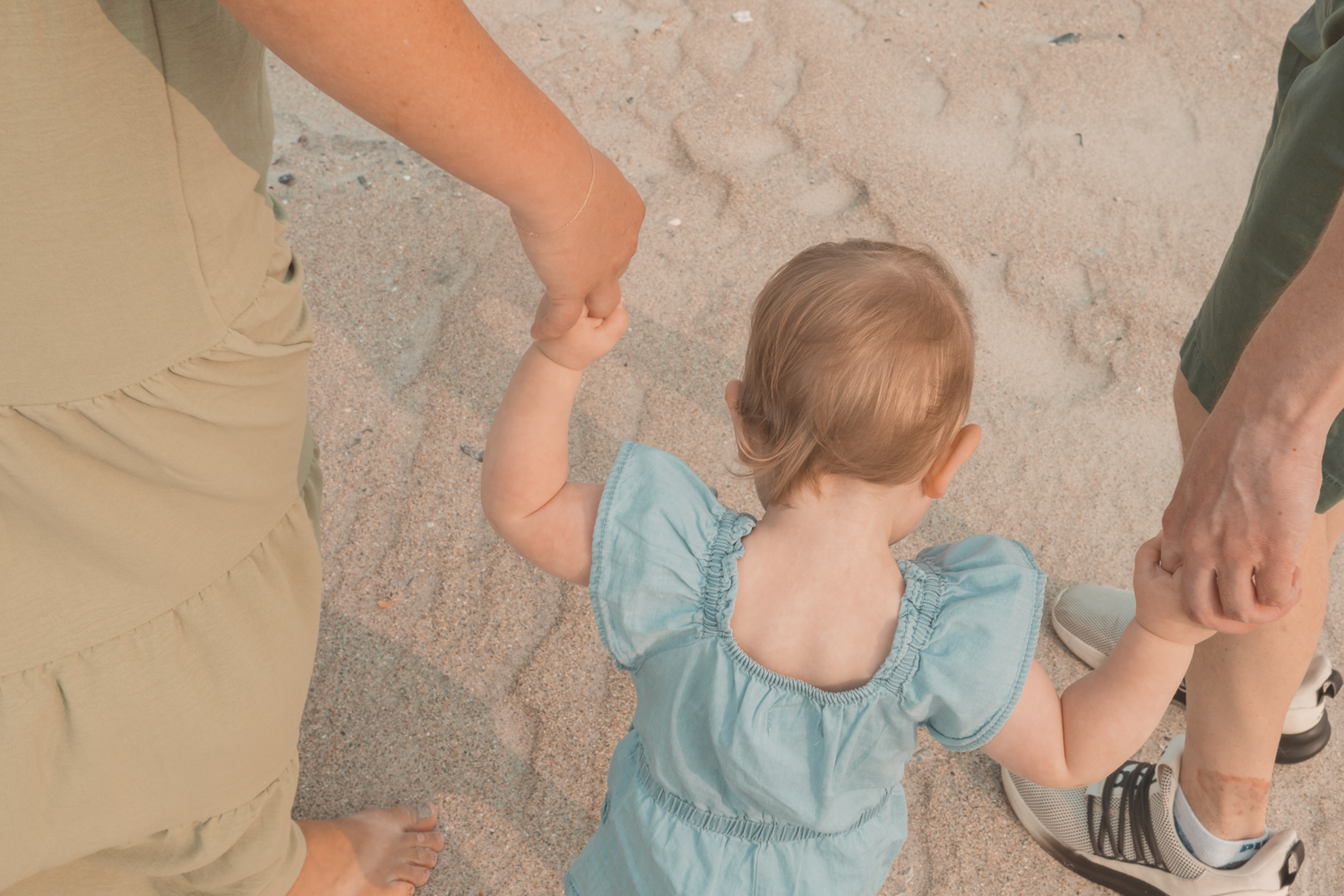 A toddler in a light blue dress holds hands with adults while walking on a textured surface at the beach.